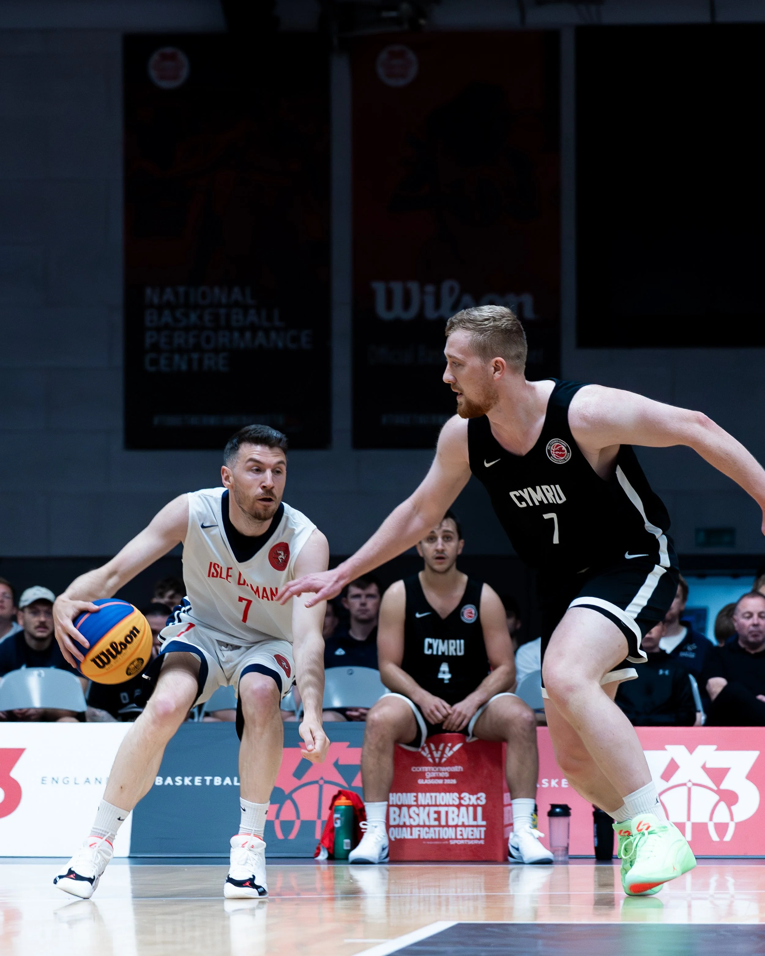 Two basketball players competing for the ball during a game. One player wearing a white jersey with red and blue accents and the number 7, and the other wearing a black jersey with the number 7 and the word 'Cymru'. Several spectators are seated behi