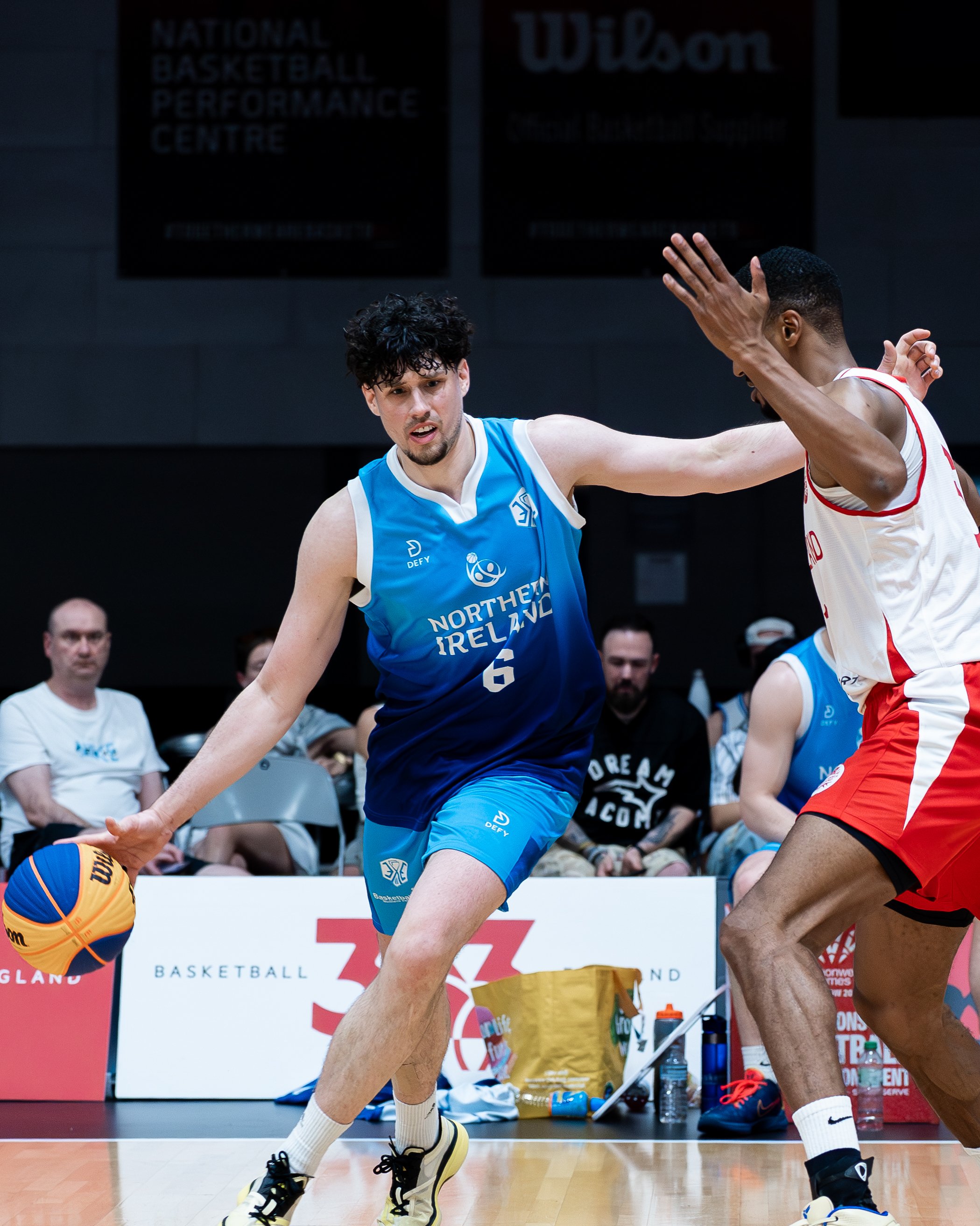 Two basketball players in a game, one in a blue jersey and the other in a white and red jersey, reaching for the ball on an indoor court, with spectators and team equipment visible in the background.