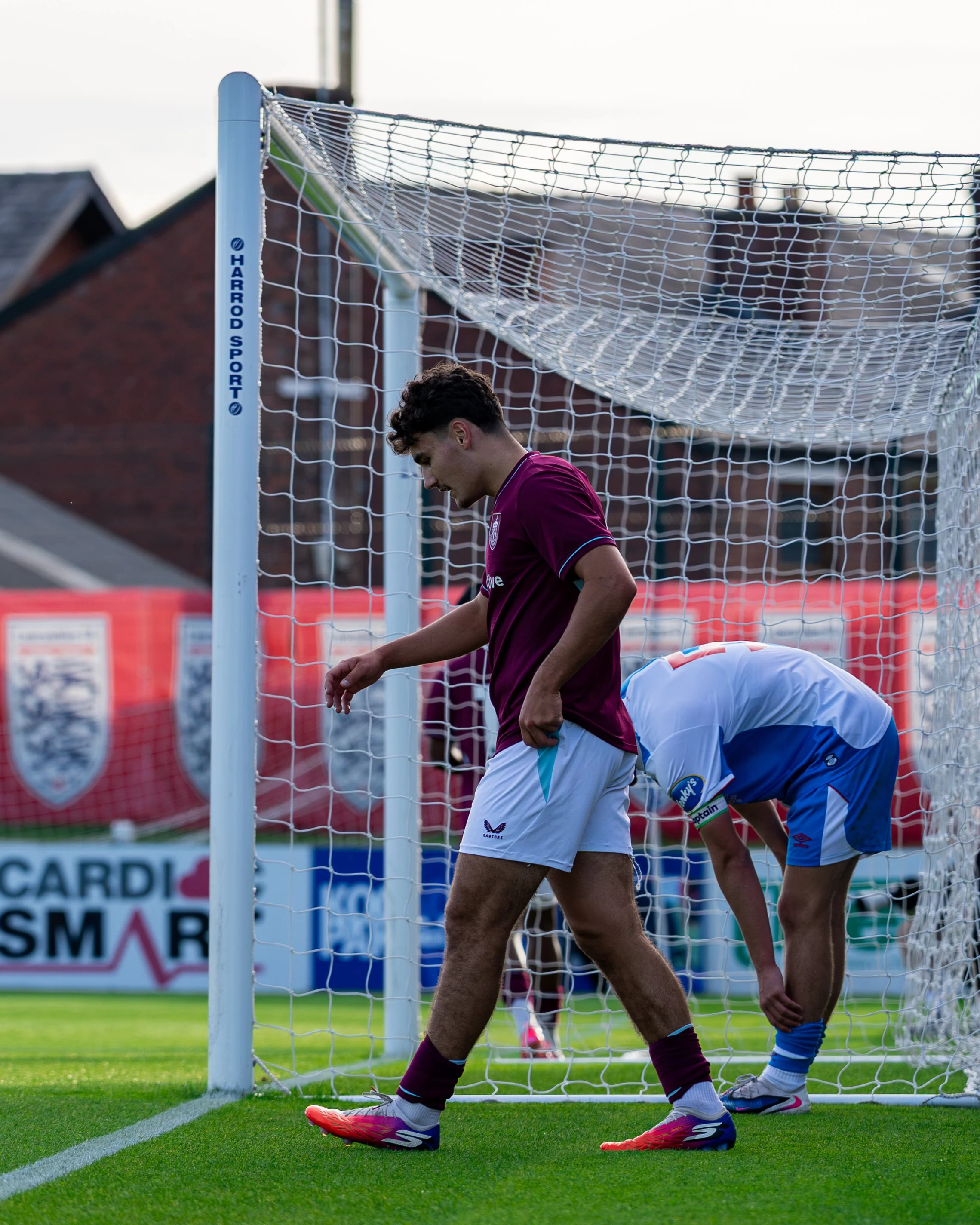 Soccer players standing near a goalpost on a field, with one player in maroon and white uniforms and another in blue uniform, during a game.