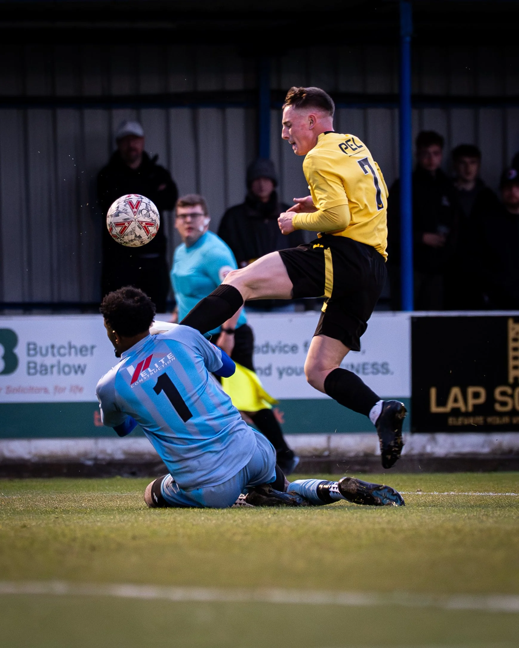 A soccer player in a yellow jersey jumps in the air as a goalkeeper in a light blue uniform slides on the ground during a match, with spectators watching in the background.