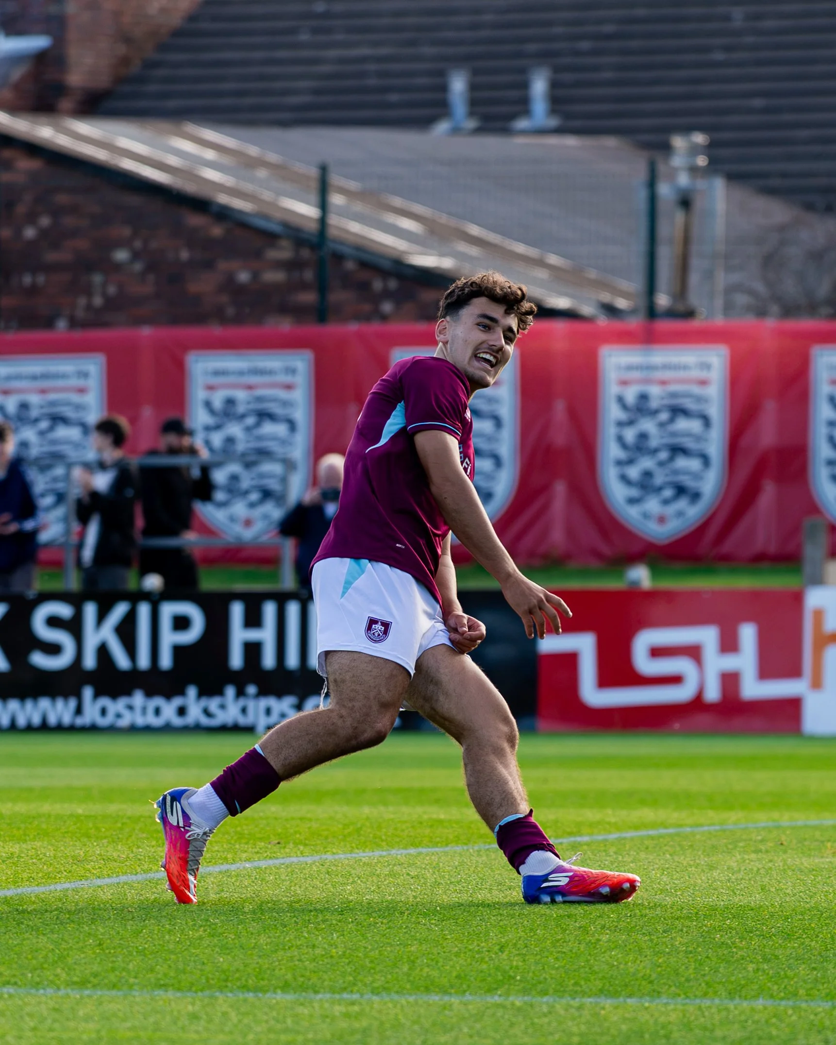 A soccer player in a maroon jersey and white shorts runs on a green field with pink cleats and purple socks, with a red advertising banner and spectators in the background.