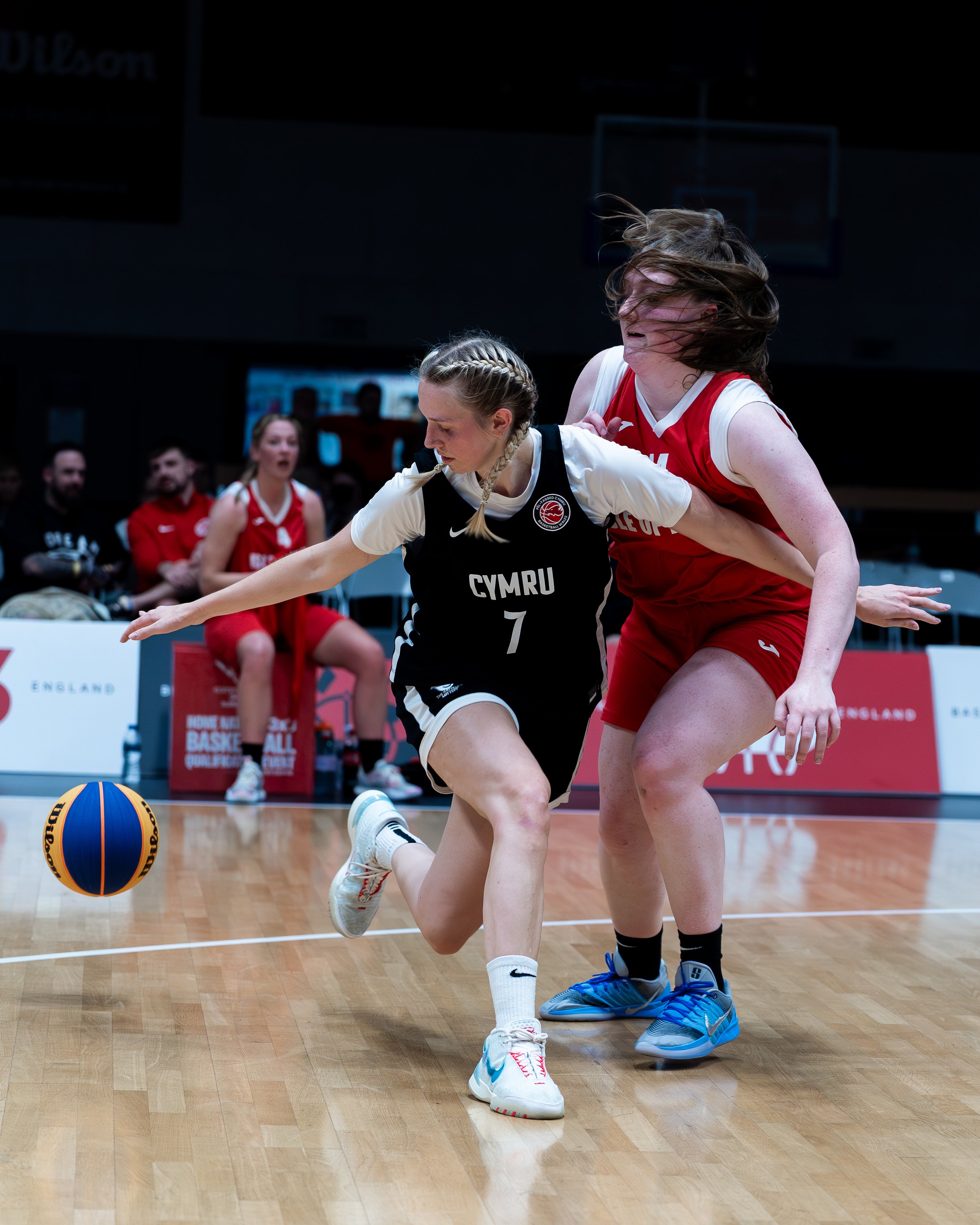 Two female basketball players competing for the ball on the court. One player in a black and white uniform with 'Cymru' and the number 7, the other in a red uniform. Crowded bench in the background.