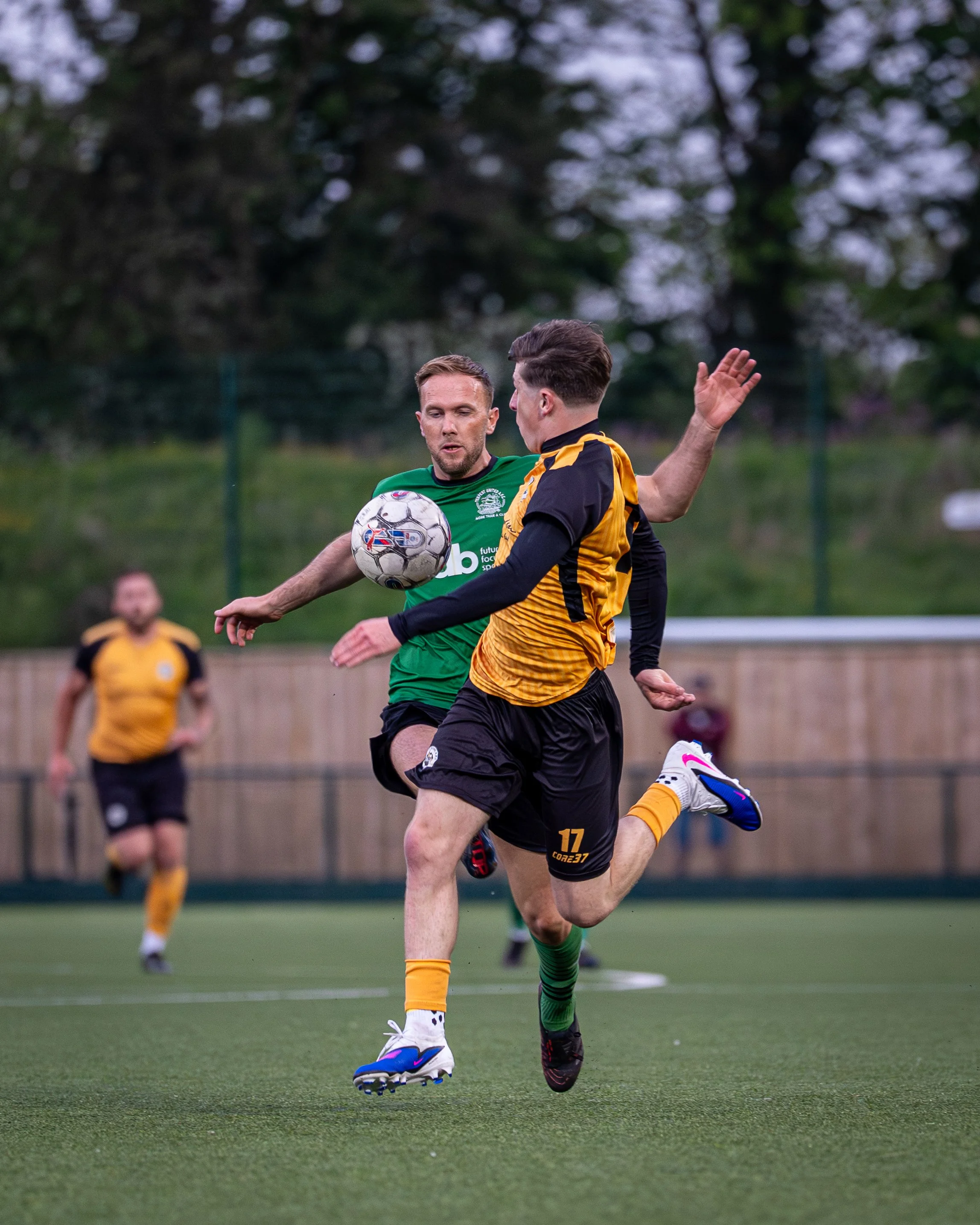 Two soccer players in action during a game on a field, one in a green jersey and the other in a yellow and black jersey, competing for control of the ball.