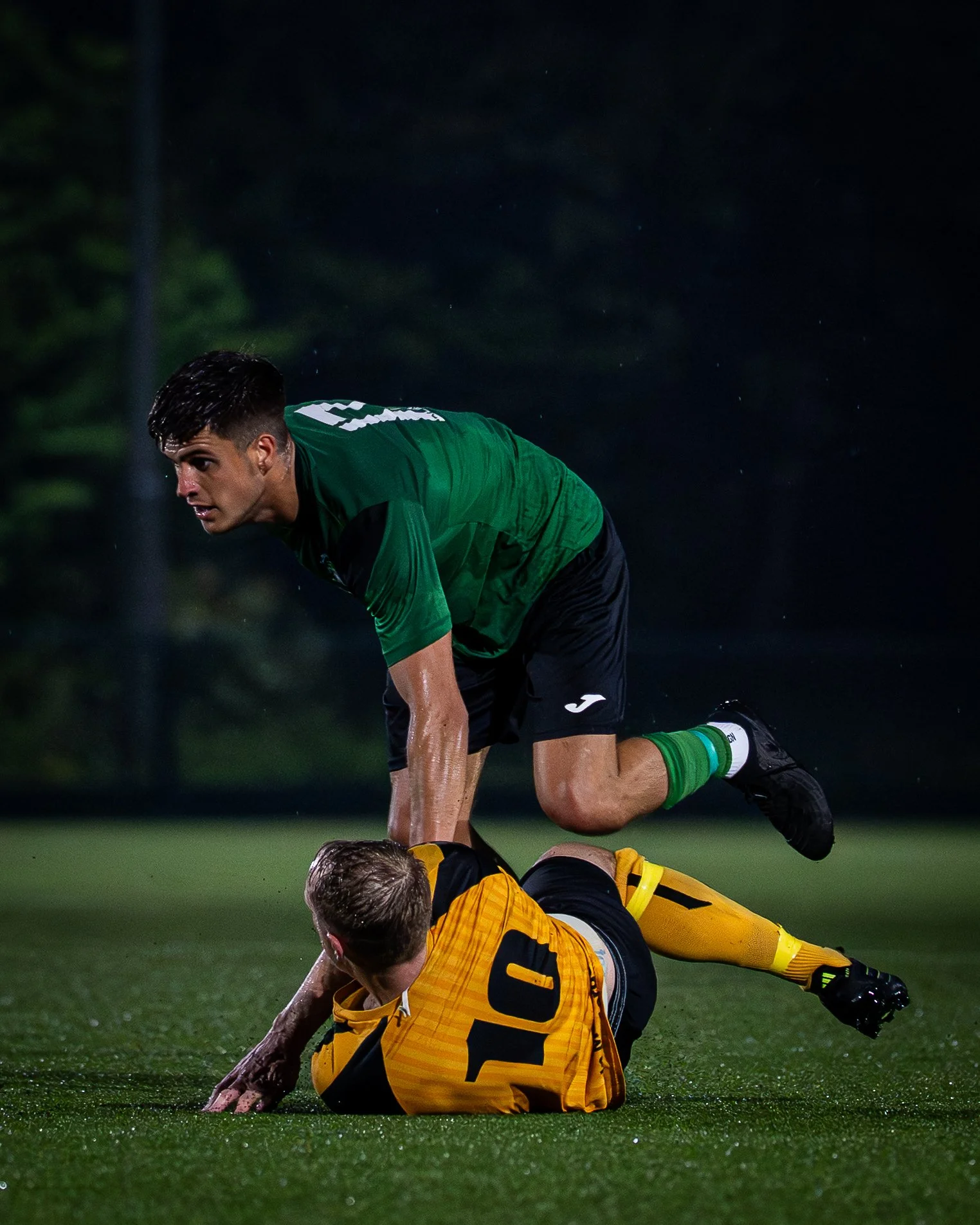 A soccer player in a green jersey is on his hands and knees on the field, looking towards another player in a yellow goalkeeper jersey who is lying on the ground. It appears to be a nighttime game.