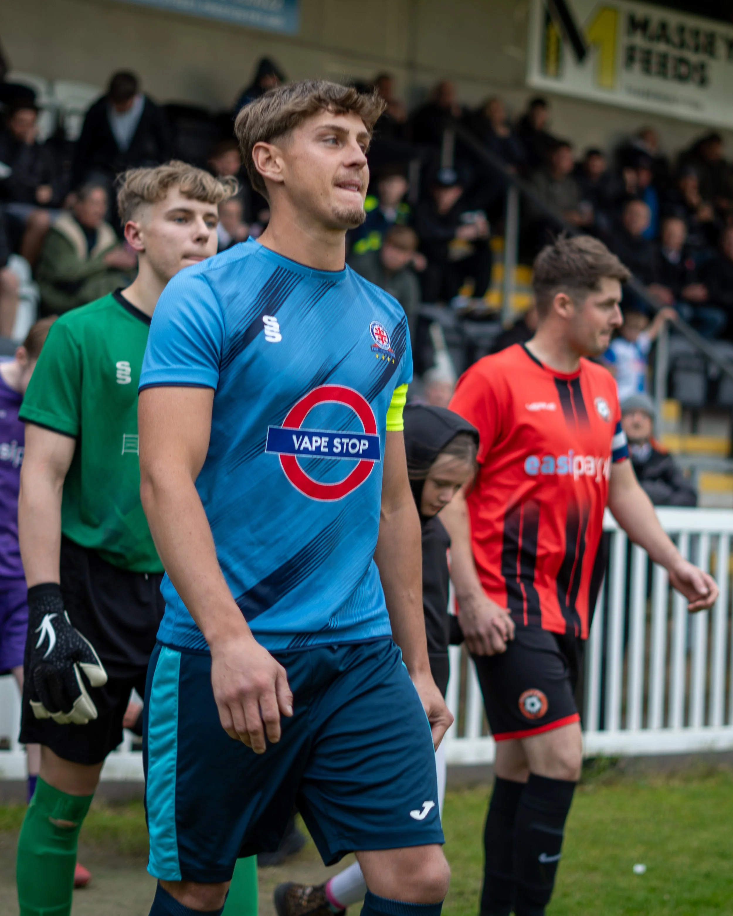Three soccer players walking on a field, with a crowd in the background. The player in front is wearing a blue jersey with a logo and a sign that says 'VAPE STOP'. The second player behind him is in a green jersey, and the third in line is in a red j