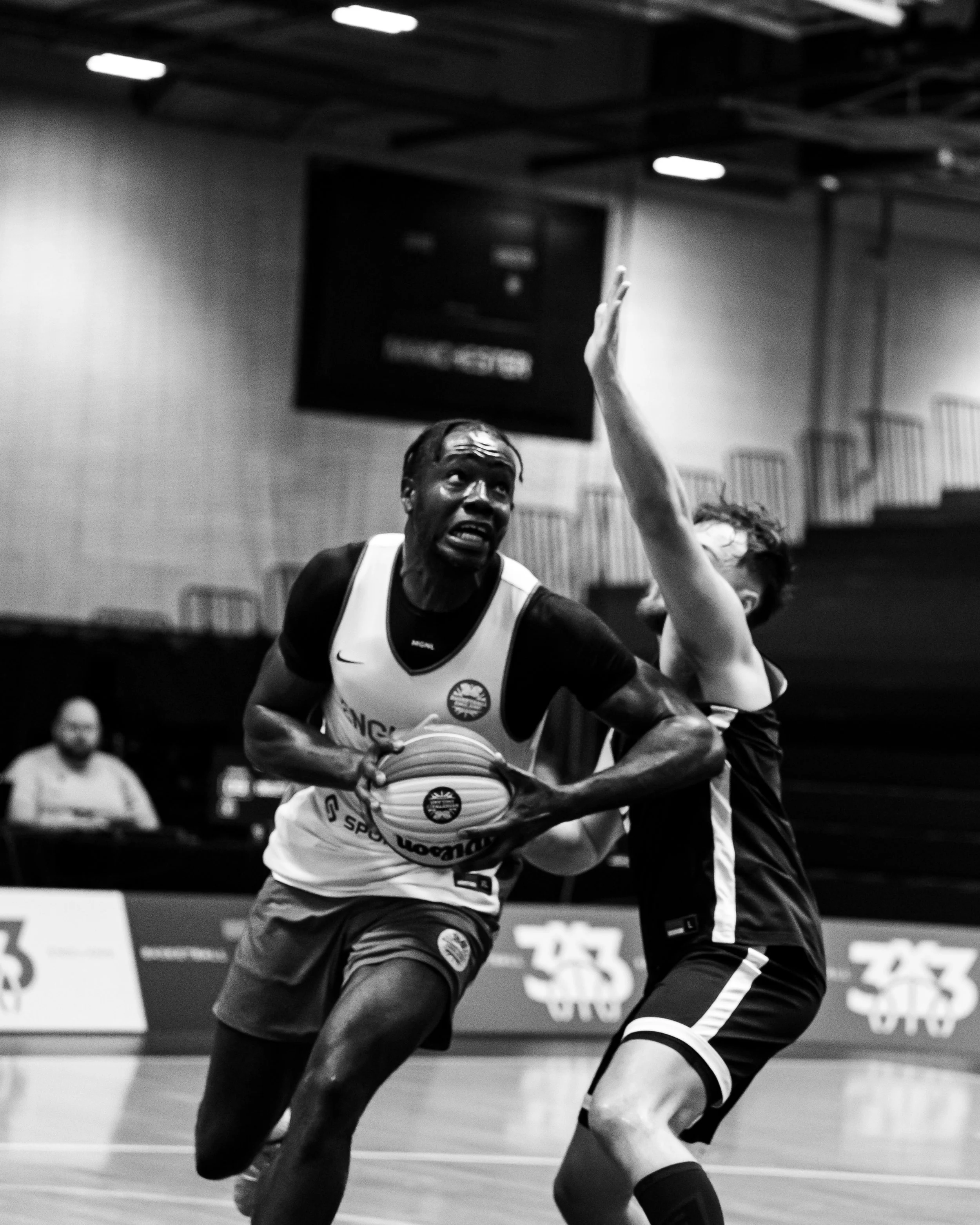 Black and white photo of two basketball players competing for possession of the ball during a game in an indoor gym. One player is wearing a jersey with the team logo and the other player is reaching over him.