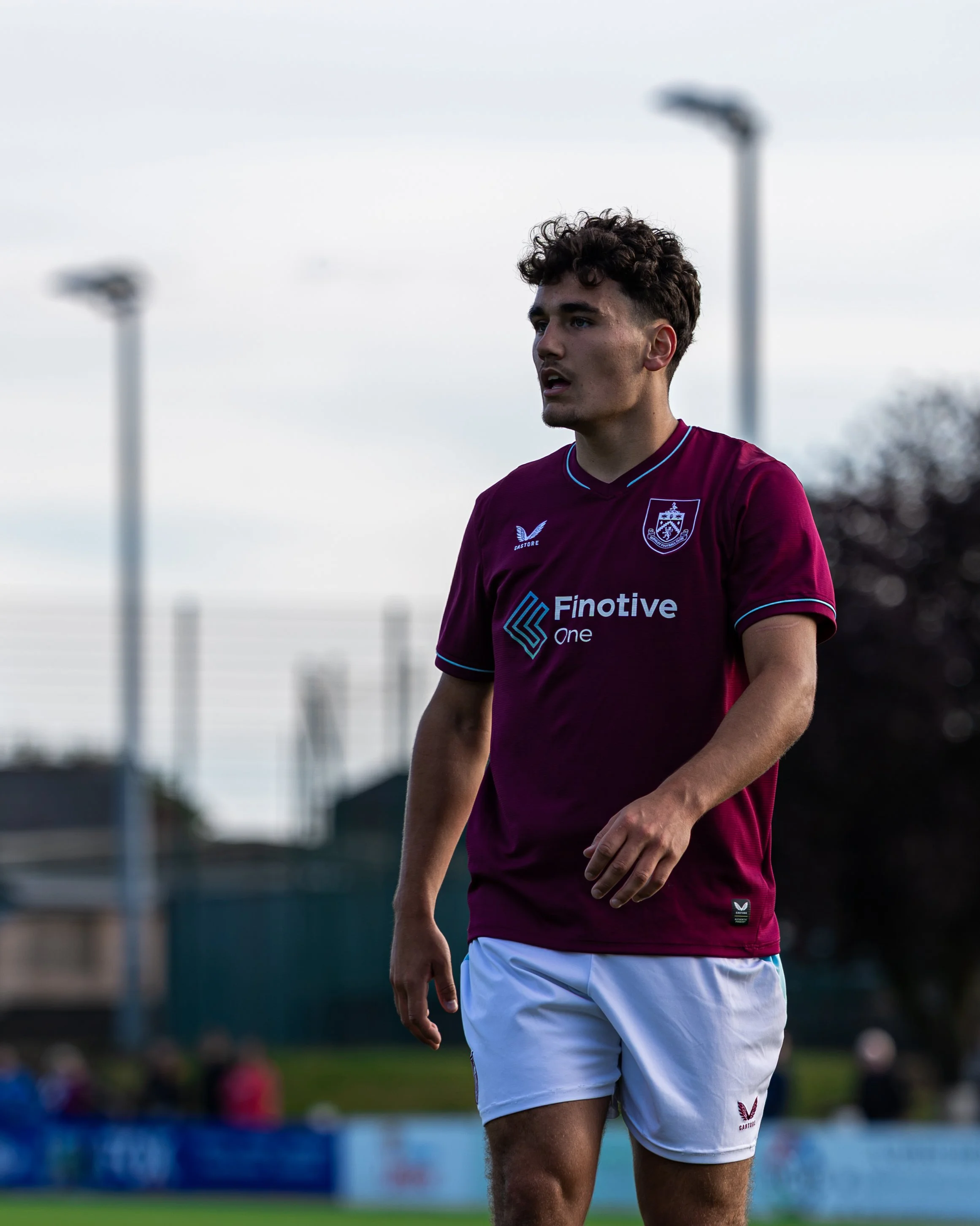 Close-up of a young male soccer player with curly hair wearing a maroon jersey and white shorts, standing on a soccer field during daytime.