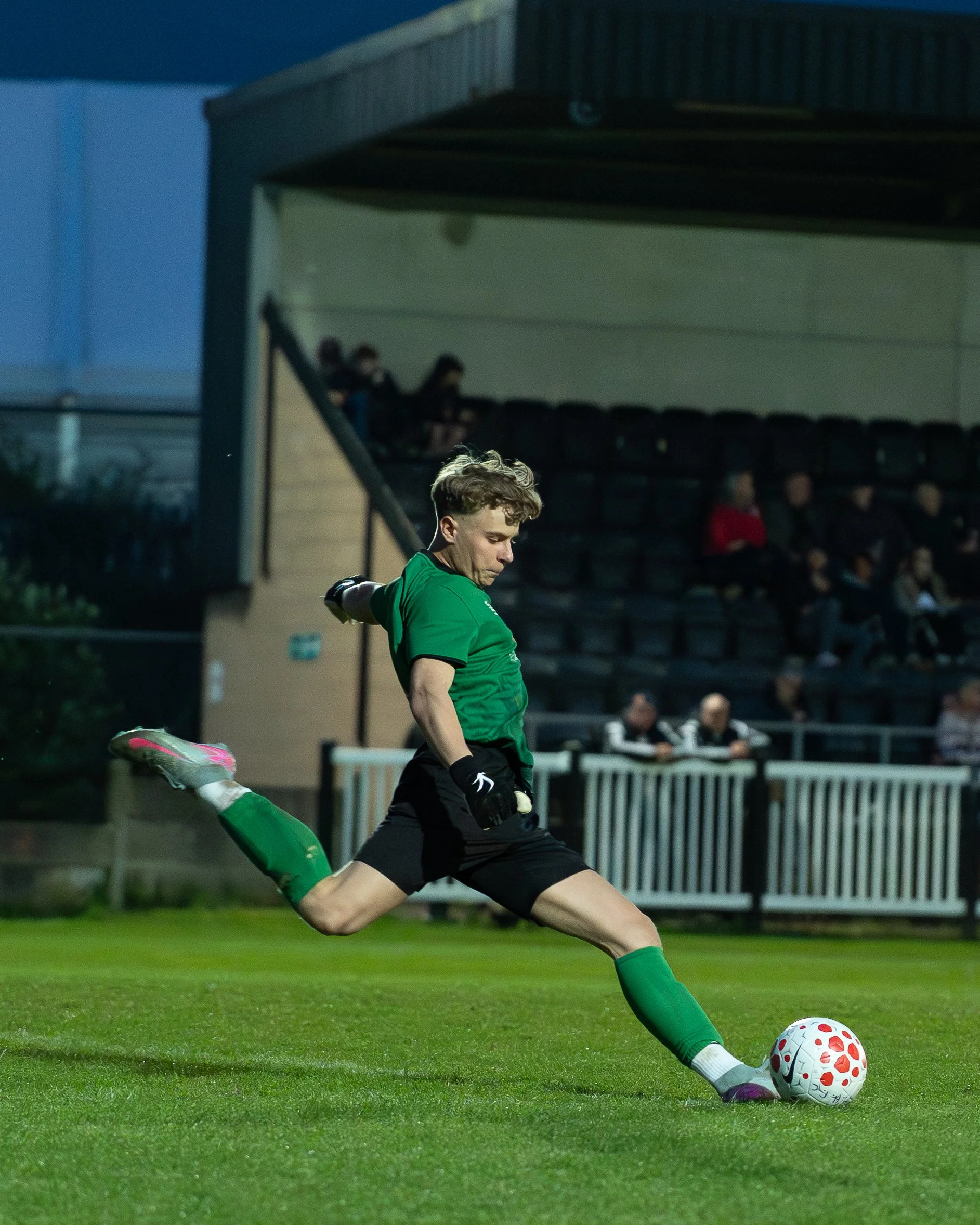 A soccer player in a green jersey and black shorts kicks a white and red soccer ball on a green field during evening, with spectators and a covered bench area in the background.