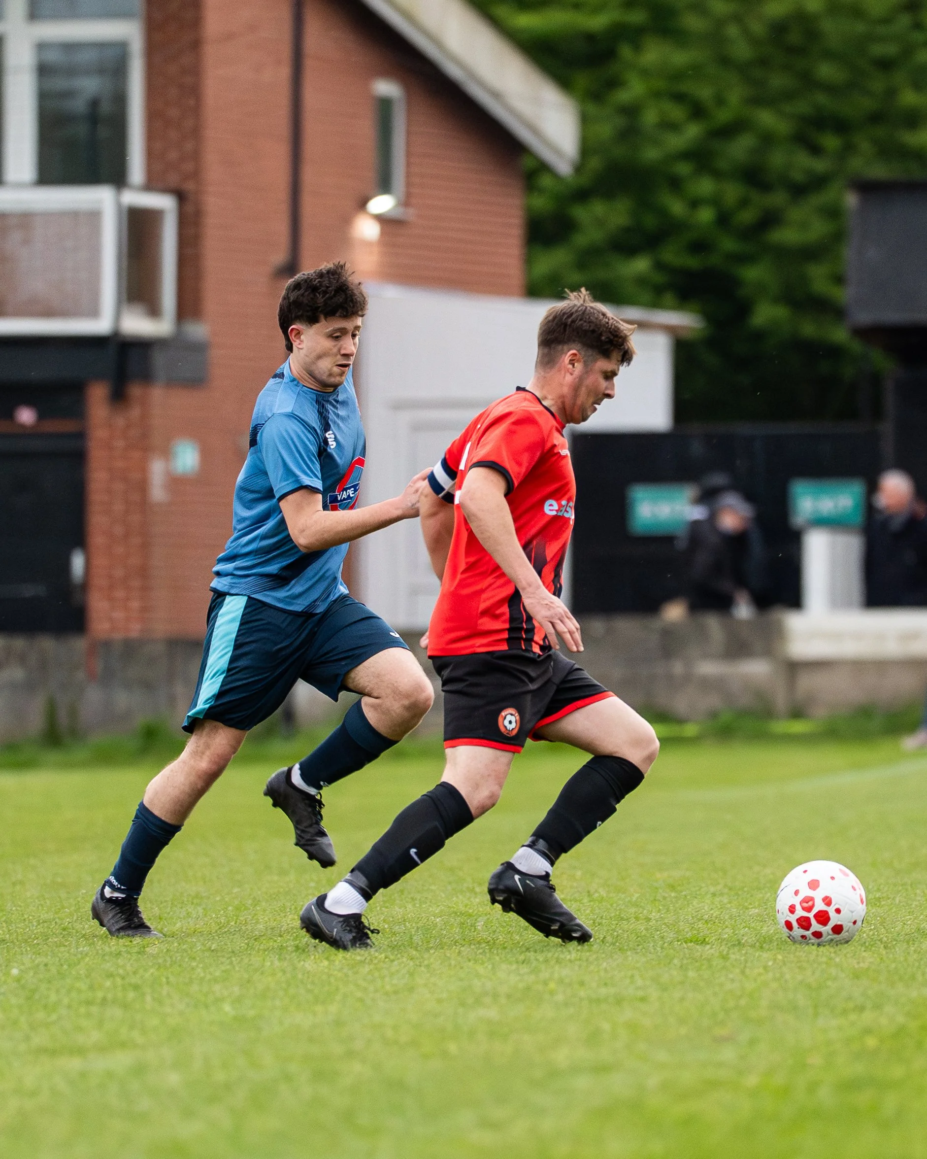 Two male soccer players competing for the ball on a green field, one wearing a blue uniform and the other wearing a red and black uniform, with a white and red soccer ball nearby.
