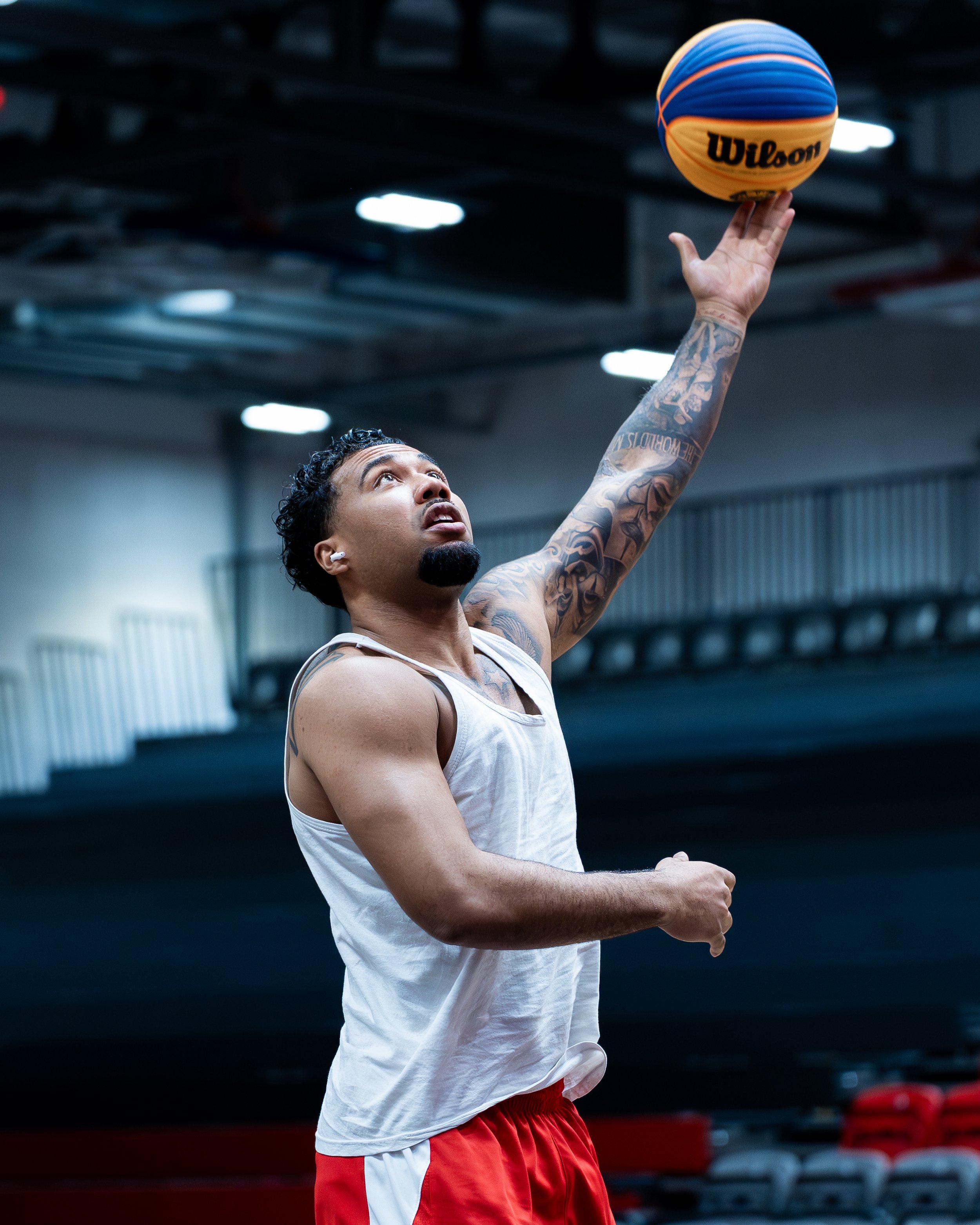 A male basketball player with tattoos on his arm and a mustache takes a shot with a Wilson basketball indoor gym.