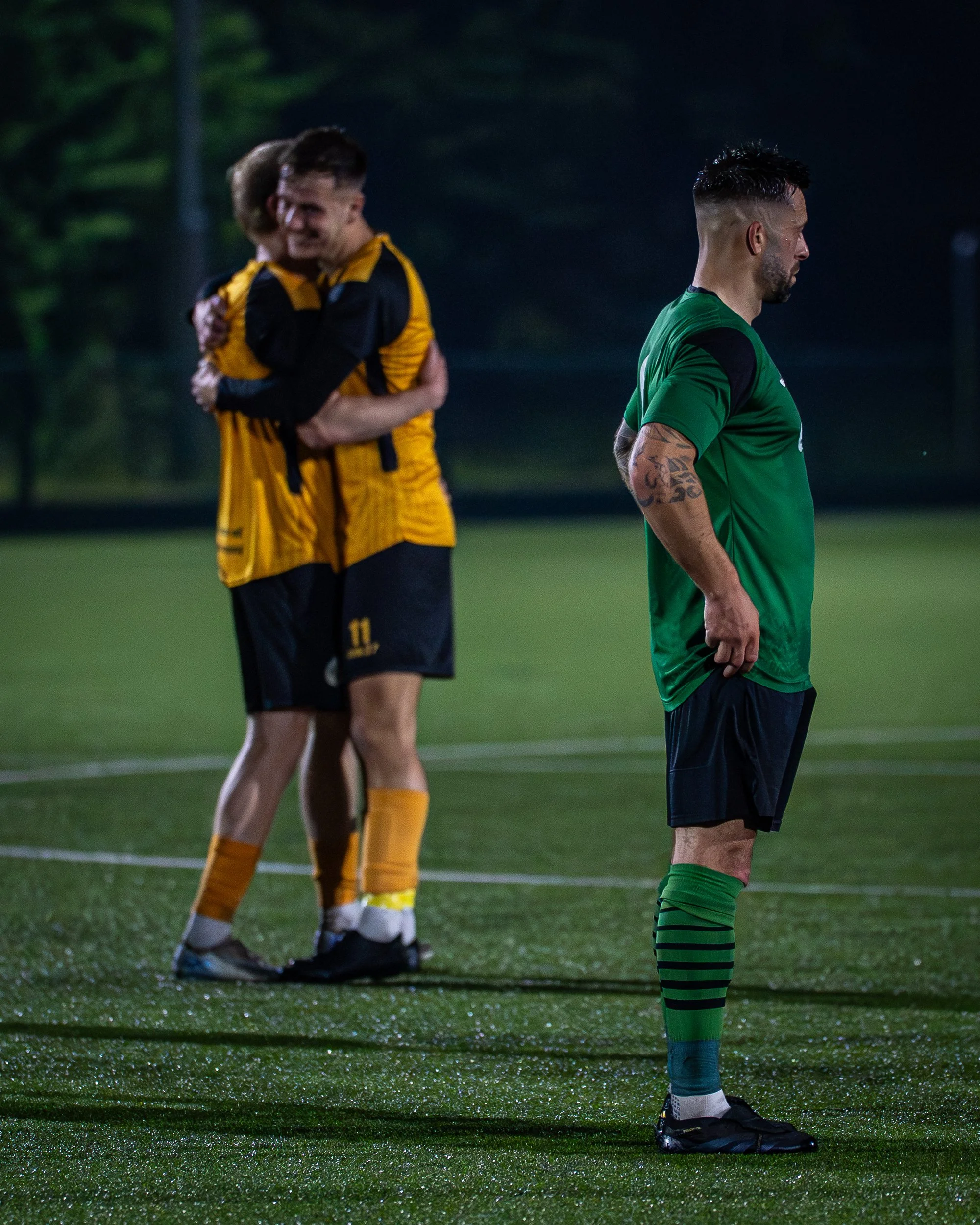 Soccer players on a field during a game at night, with one player standing with arms crossed and two others embracing in the background.