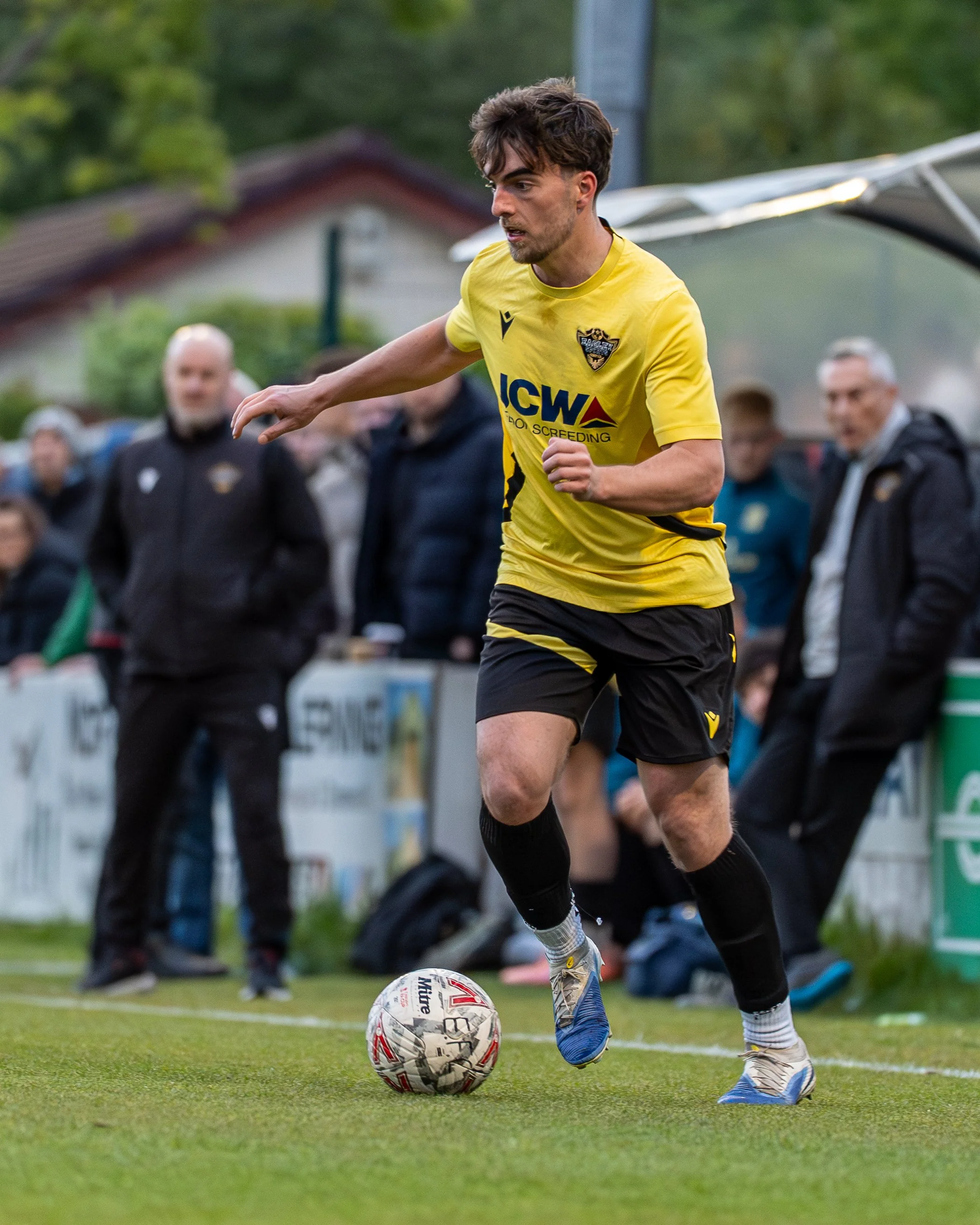 A young male soccer player in a yellow jersey and black shorts kicks a soccer ball on a field, with spectators and team staff watching in the background.