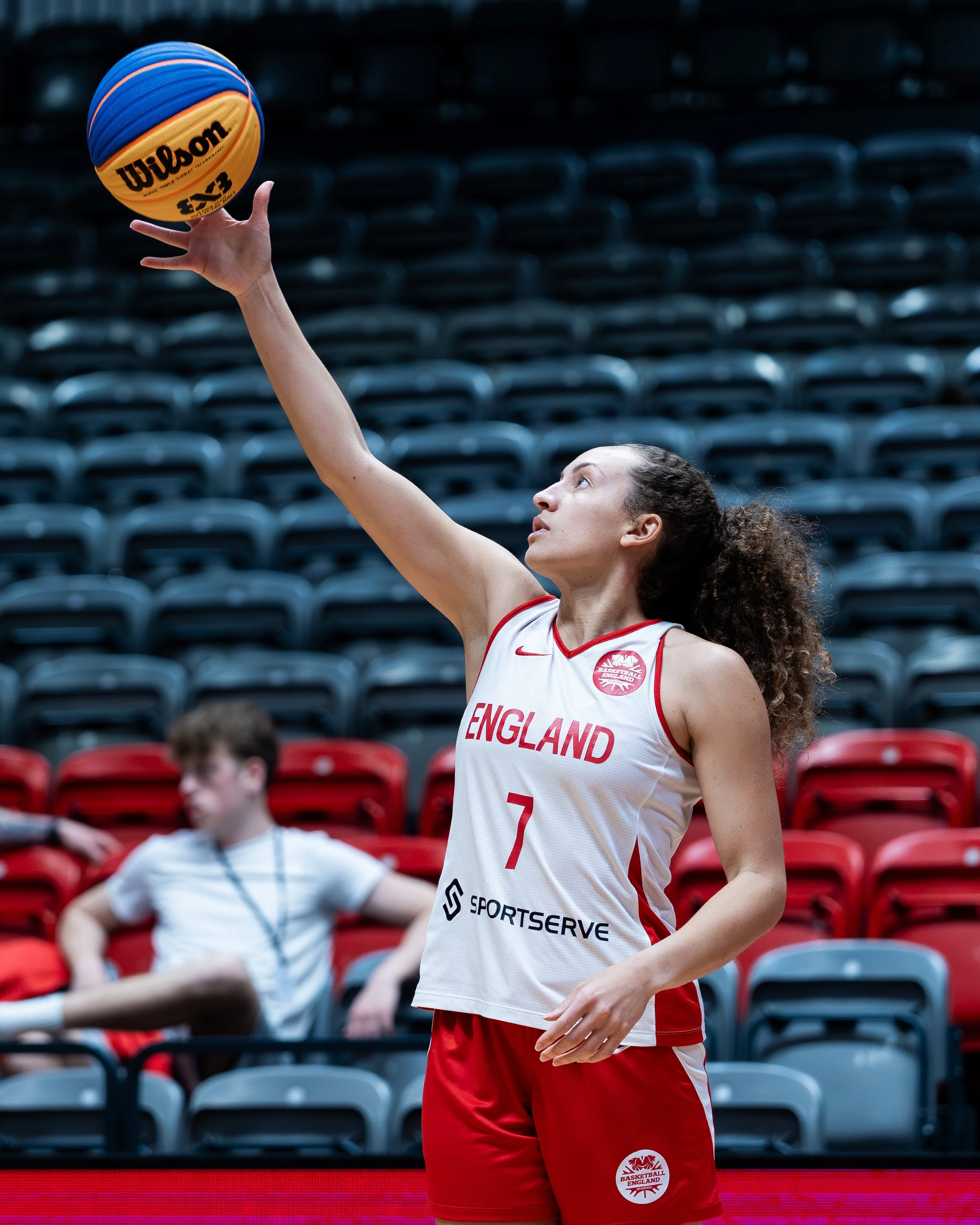 A female basketball player with curly hair and light skin, wearing a white and red England team jersey, is practicing shooting a basketball on an indoor court with empty spectator seats in the background.