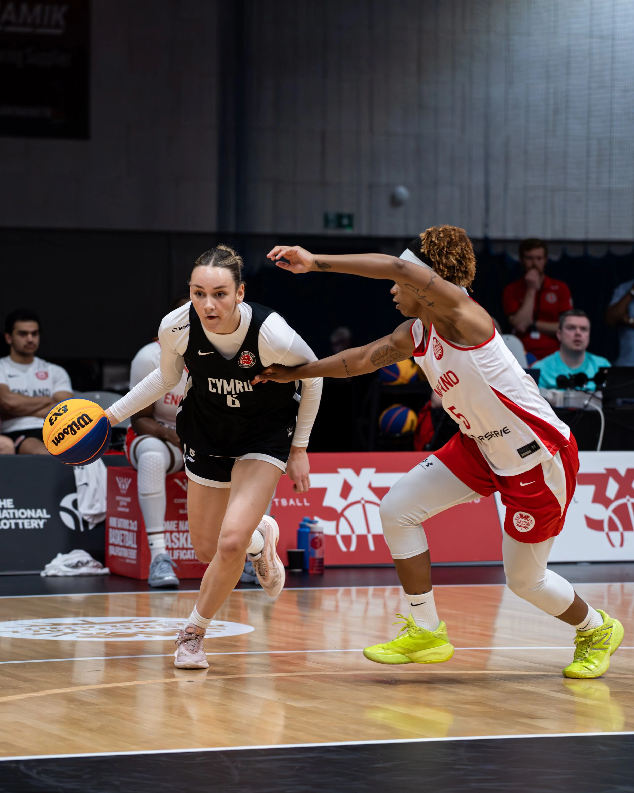 Two female basketball players from opposing teams compete for possession of the basketball during a game. One player wears a white and red uniform, and the other wears a black and white uniform. They are on a wooden basketball court with spectators a
