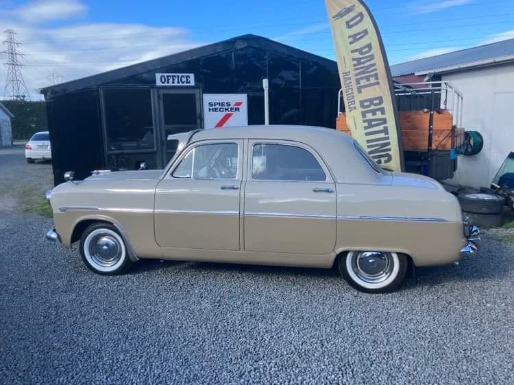 A vintage beige sedan parked outside a black building with signs, one reading 'OFFICE' and another with partially visible text. There is a yellow flag with black text, likely advertising a business, and some tires stacked nearby.
