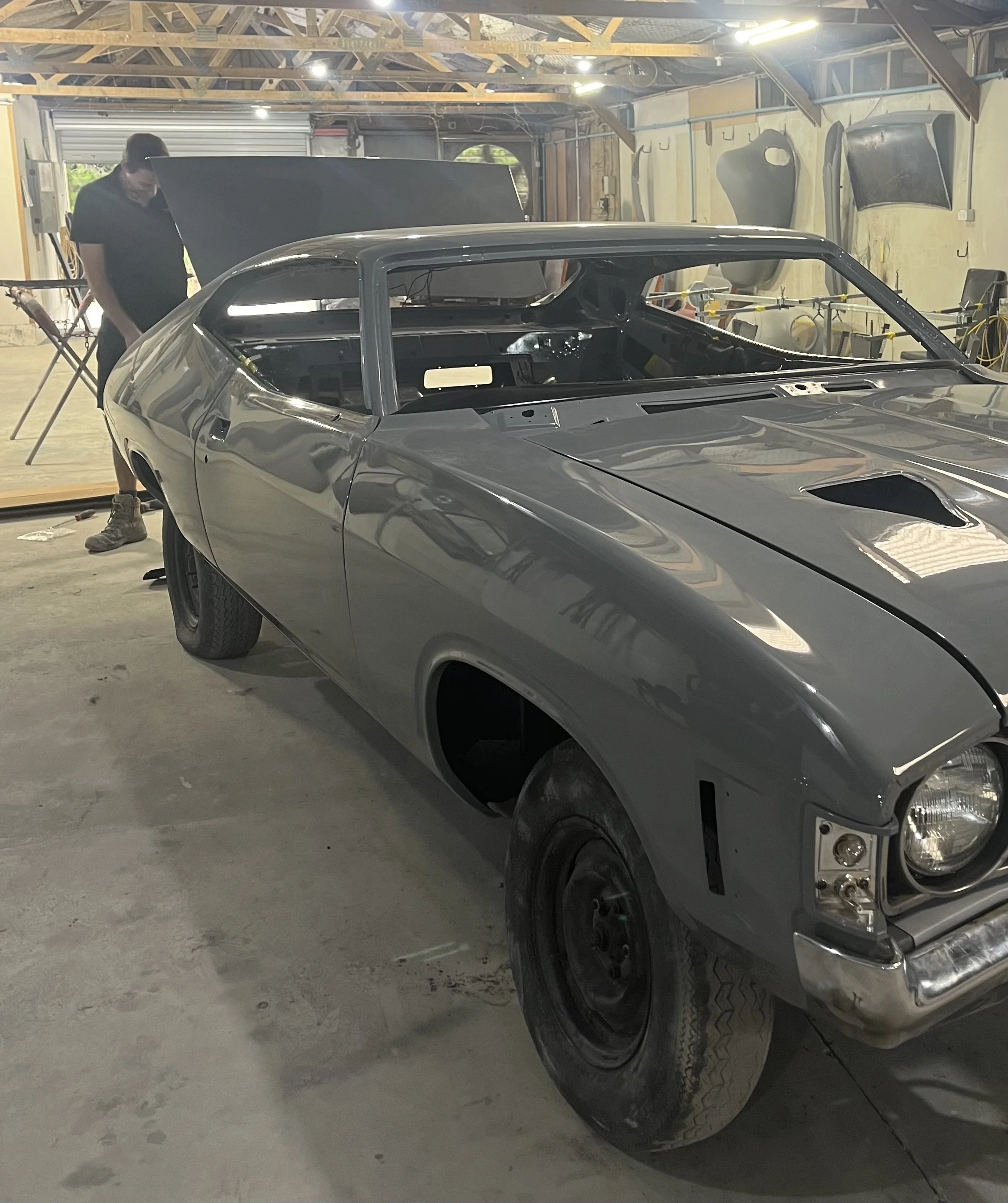A person working on a partially assembled or repaired gray car inside a garage. The garage has various car parts hanging on the wall and tools scattered around.