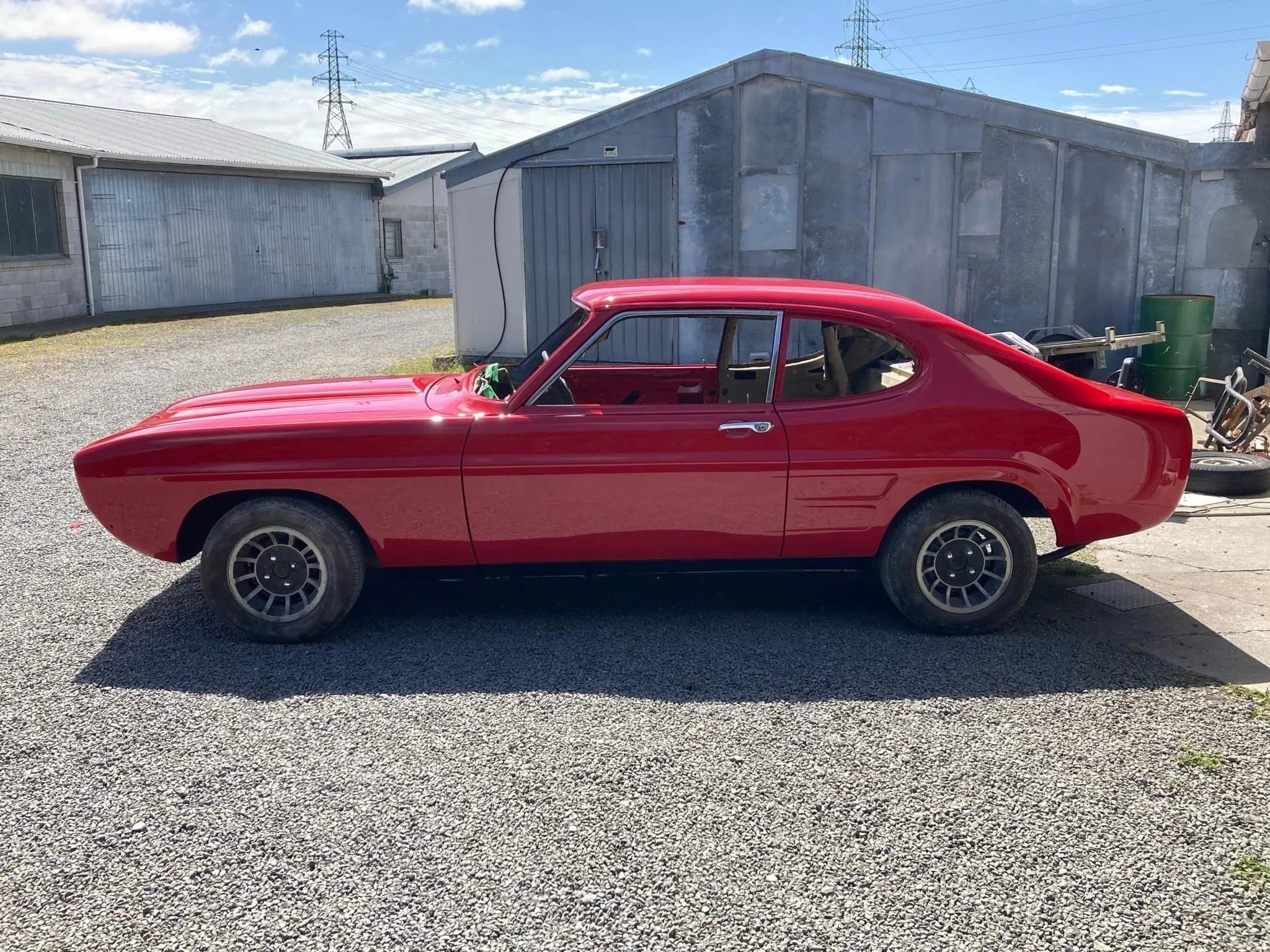 A red classic car with no windows parked outdoors on gravel, with a background of industrial-looking buildings, a green barrel, and power lines and pylons under a partly cloudy sky.