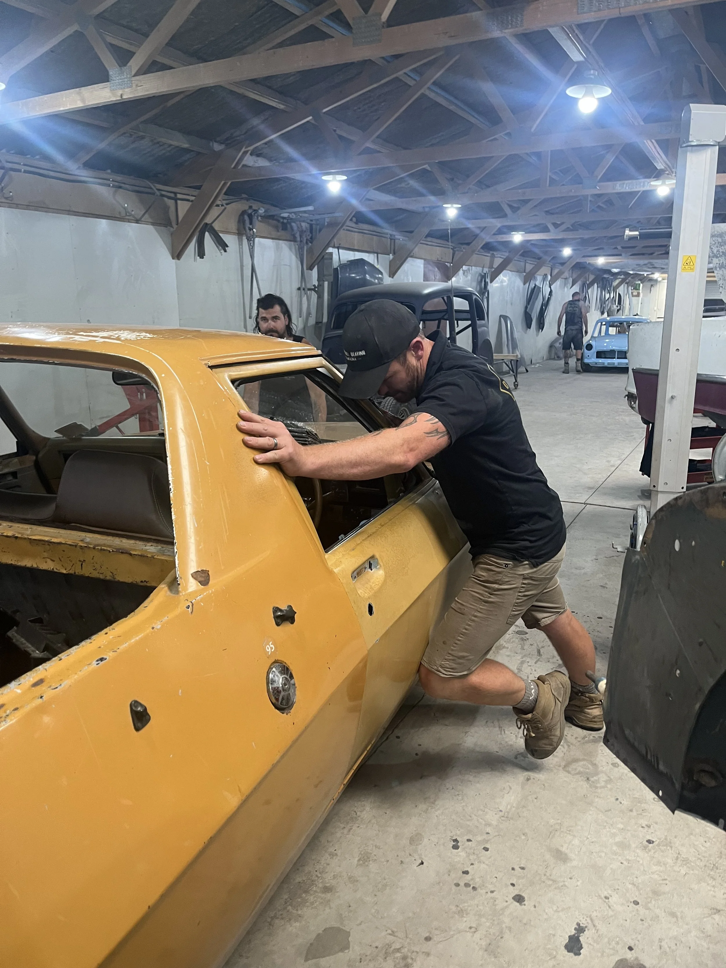 A man wearing a black cap, black t-shirt, and khaki shorts is working on a yellow vintage car in a garage or workshop. Another man in the background, with long hair and a beard, is observing. The garage has exposed wooden beams ceiling, various car parts hanging, and several old cars, including a blue race car and a black car frame. Bright overhead lights illuminate the scene.