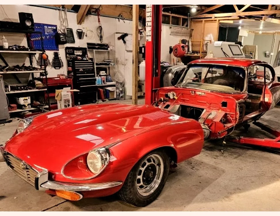 A red classic car in a garage, partially disassembled with the hood, front fenders, and headlights intact. The rear chassis and interior are exposed and it appears to be undergoing restoration or repair.