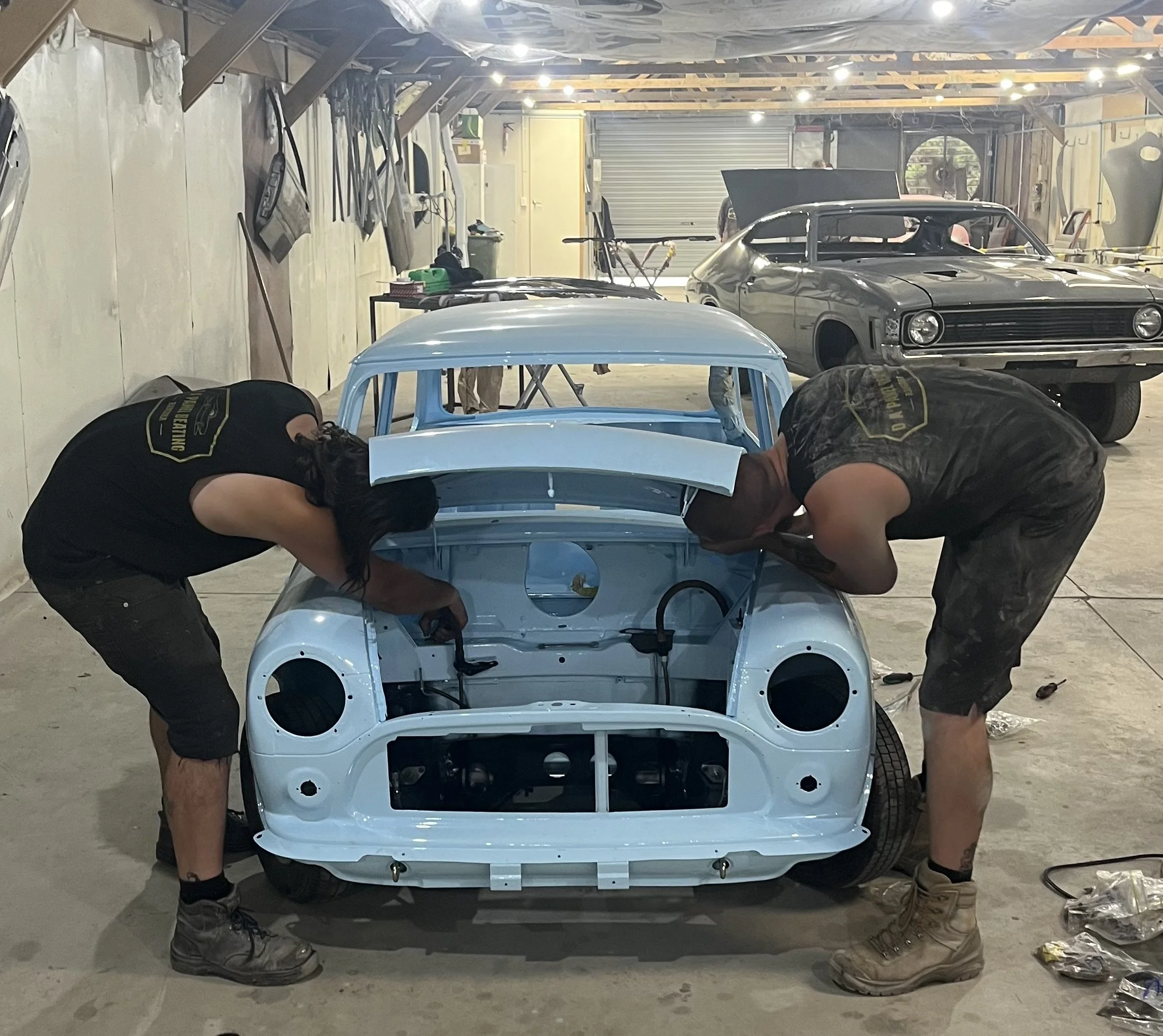 Two people working on assembling a small light blue vintage-style car inside a workshop or garage. The car's front end is partially assembled, with the body in place but no headlights or grille installed yet. One person is wearing black clothing with a badge on the back, and the other is in camouflage shorts and boots. The workshop has unfinished walls, various tools, and car parts visible around.