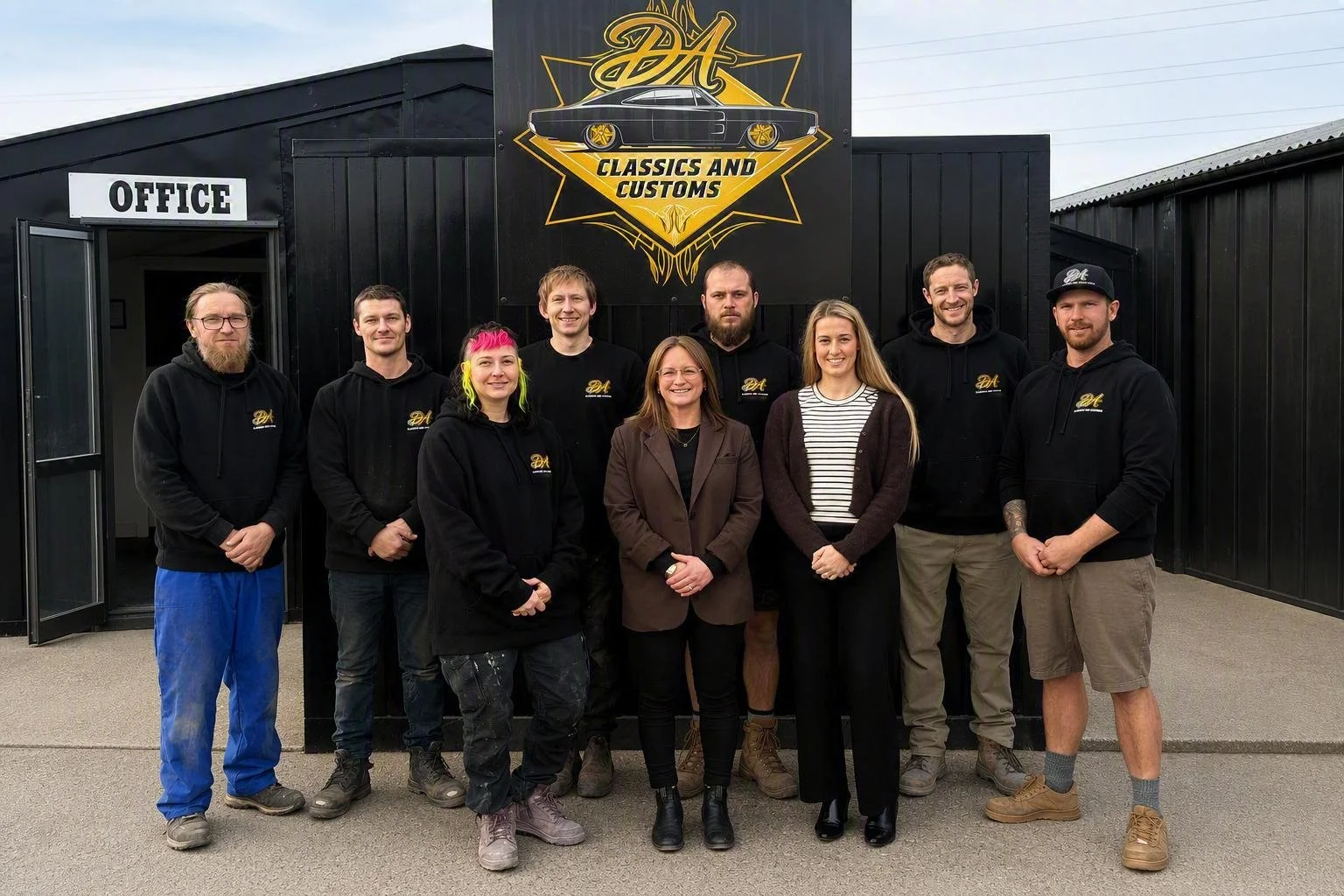 Group of nine people standing in front of a black building with a sign that reads 'Classics and Customs' and a logo featuring a classic car. An office entrance is on the left side.
