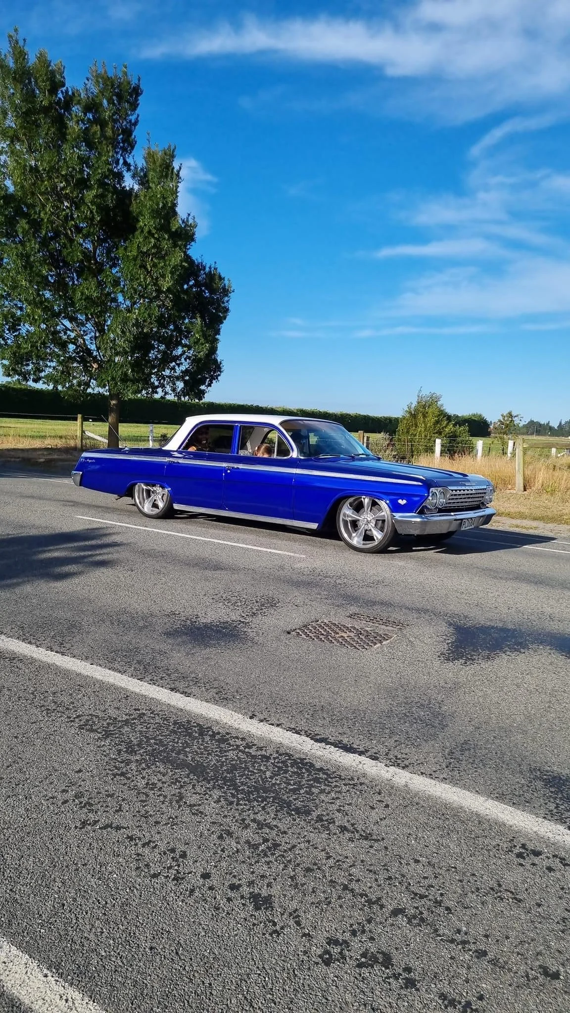 A vintage blue car with custom wheels parked on the side of a rural road under a clear blue sky with some clouds.