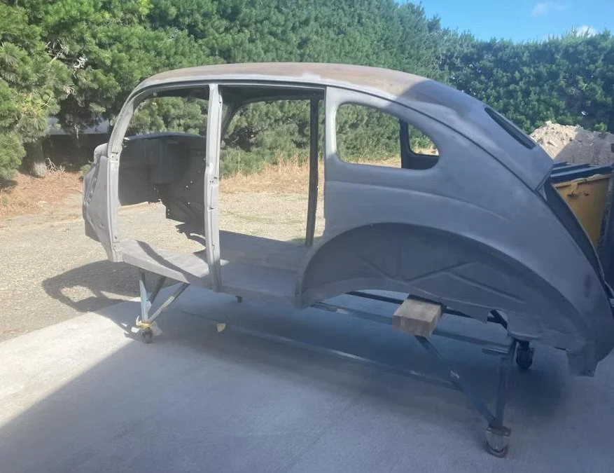 A vintage car body shell mounted on a support frame outdoors, with trees and a blue sky in the background.