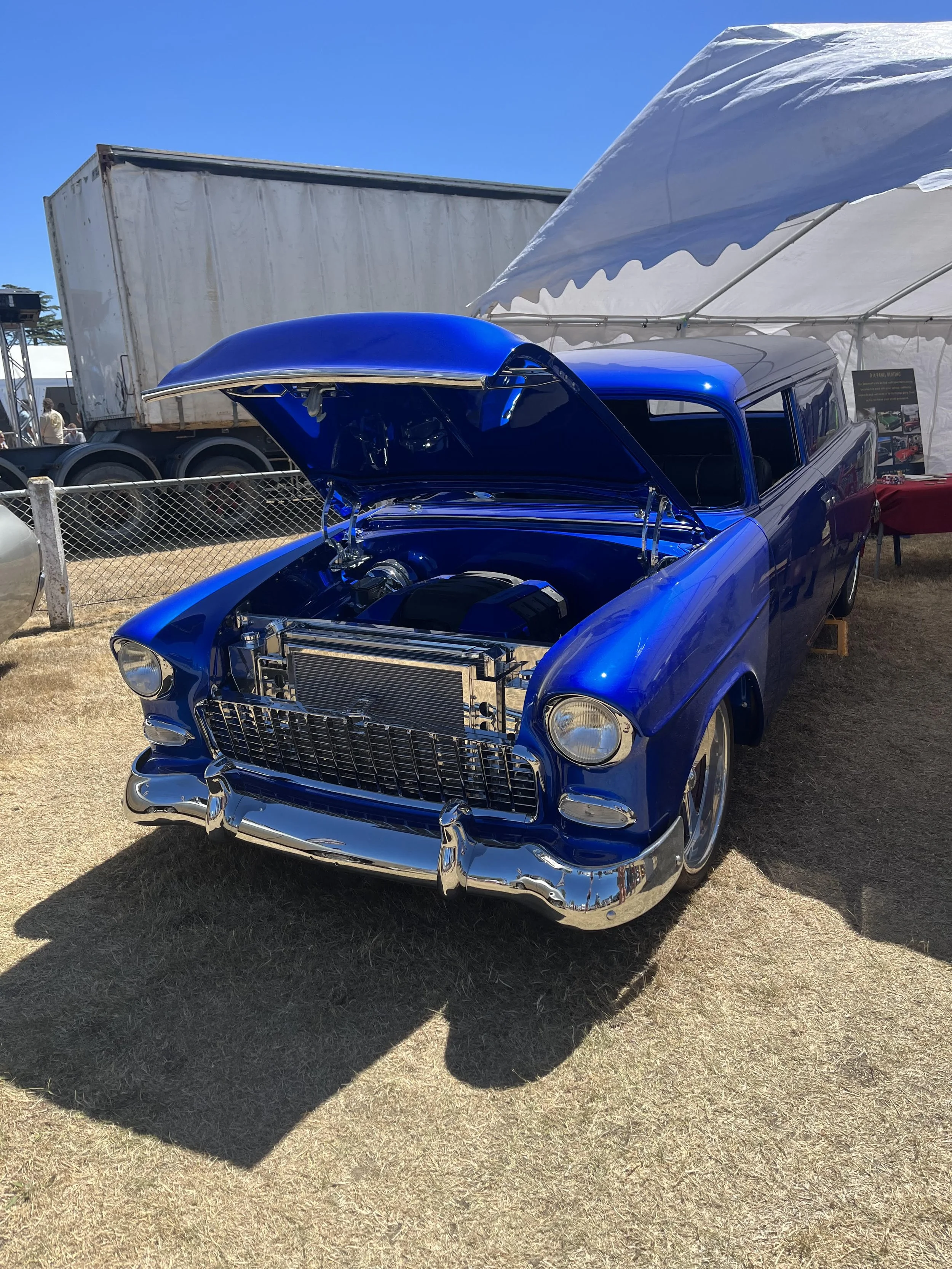 A shiny blue vintage car with its hood open, displayed at an outdoor car show event under a white tent against a clear blue sky.