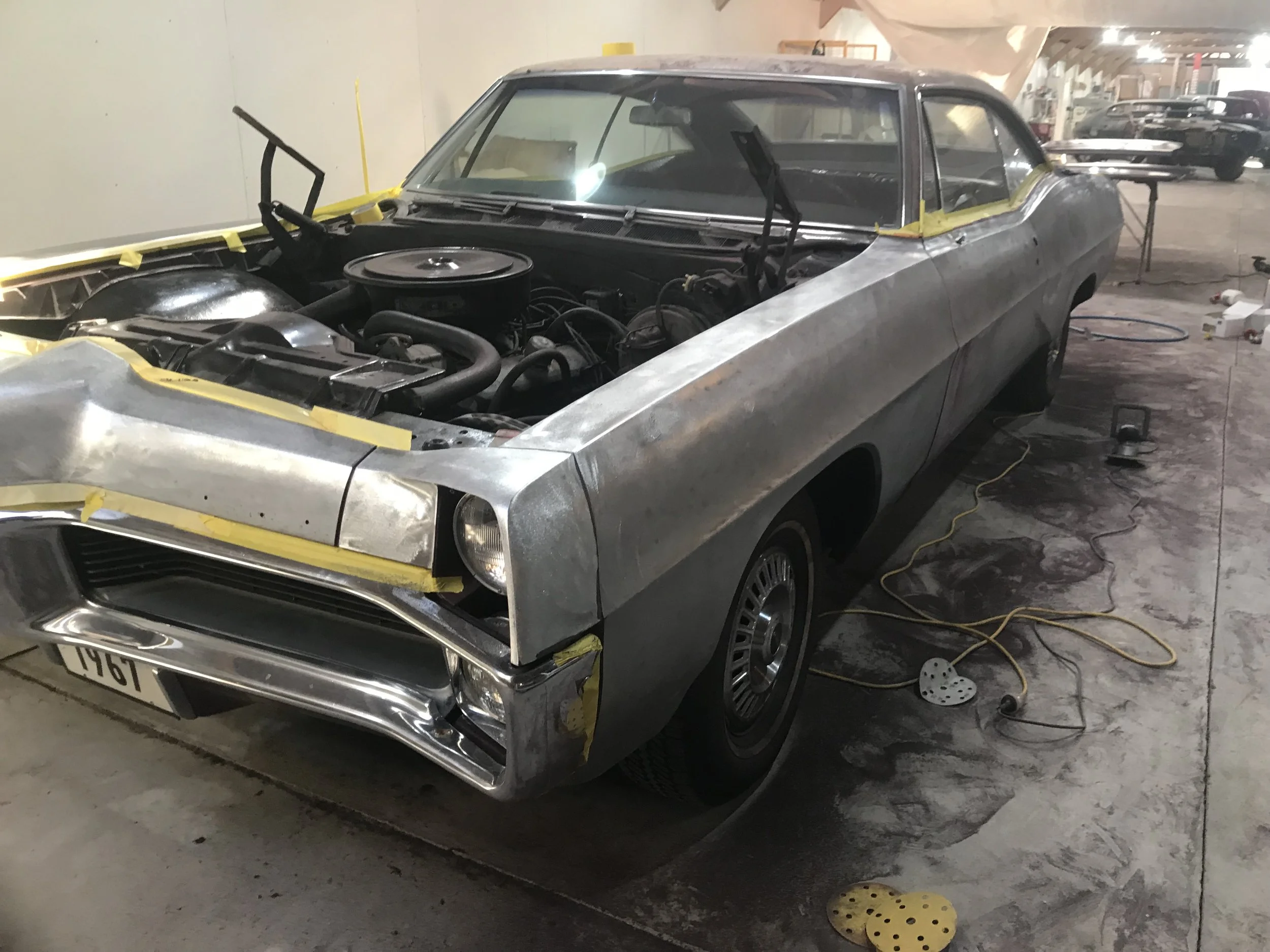 A vintage car being restored in a garage, with its hood open and parts on the floor.