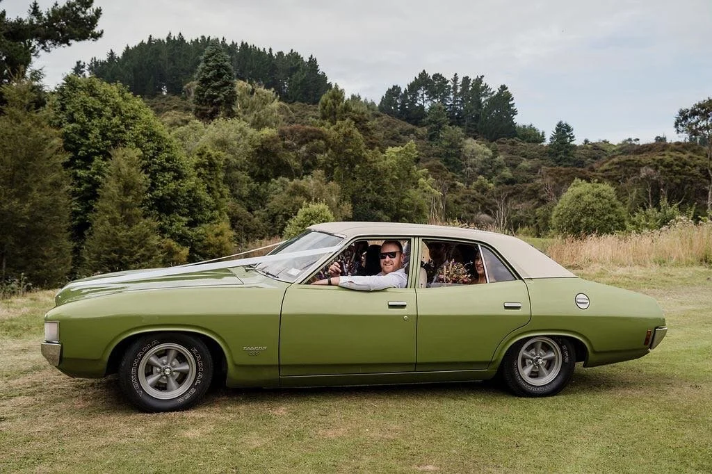 A vintage green car with a white roof parked on grass in a natural, wooded setting. There are people inside the car, with one man visible smiling and wearing sunglasses.