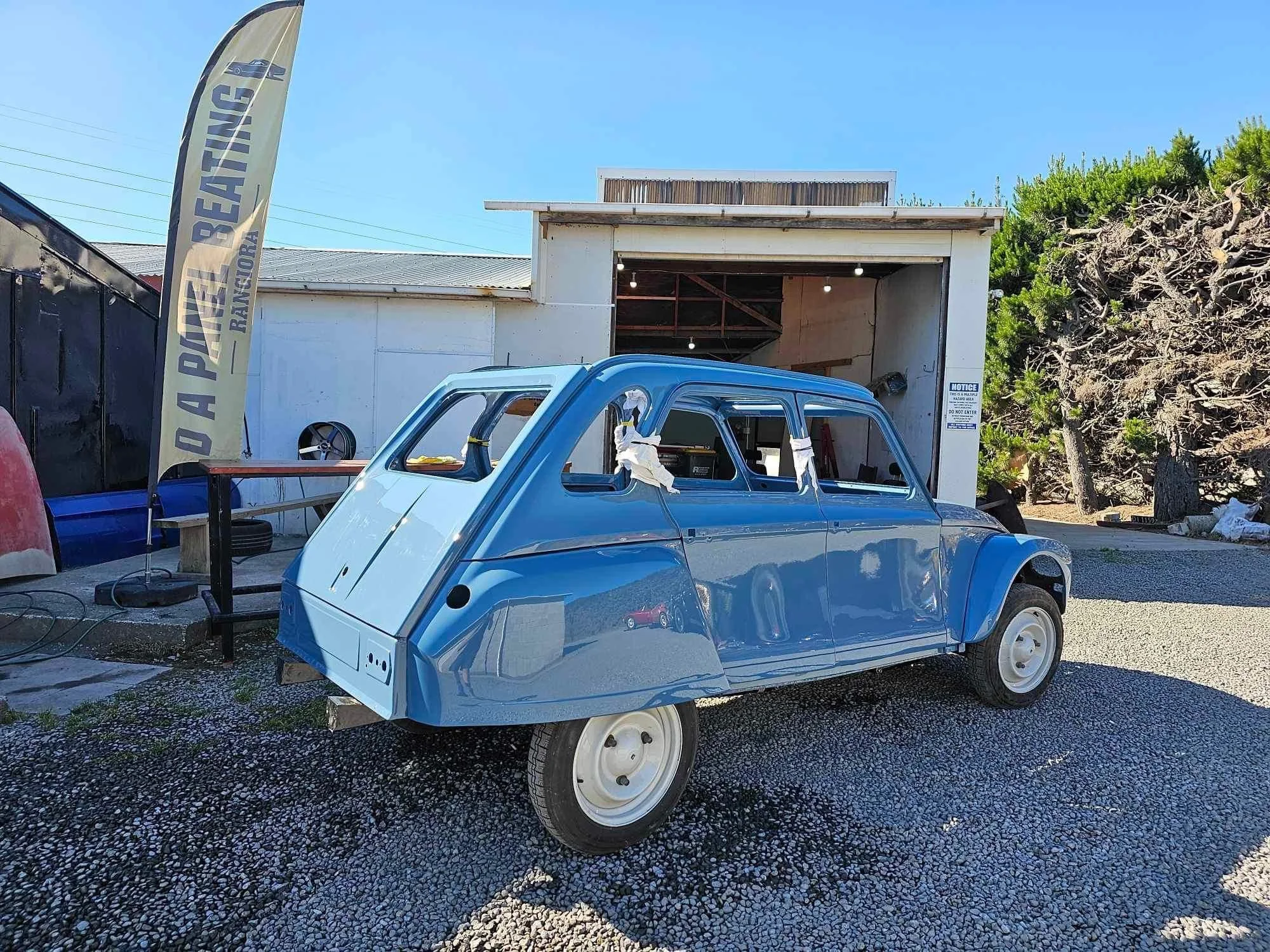 A small, unassembled blue car body in a car workshop, with a white building and a yellow flag with the words 'AUTO PAINT & BODY REPAIRS' written on it. The workshop has a gravel ground with some tools and a black fence on the left side.