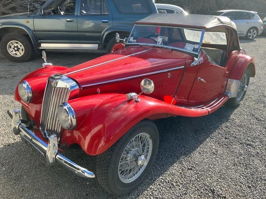 A vintage red convertible car with a soft top parked on a gravel lot, with other vehicles in the background.