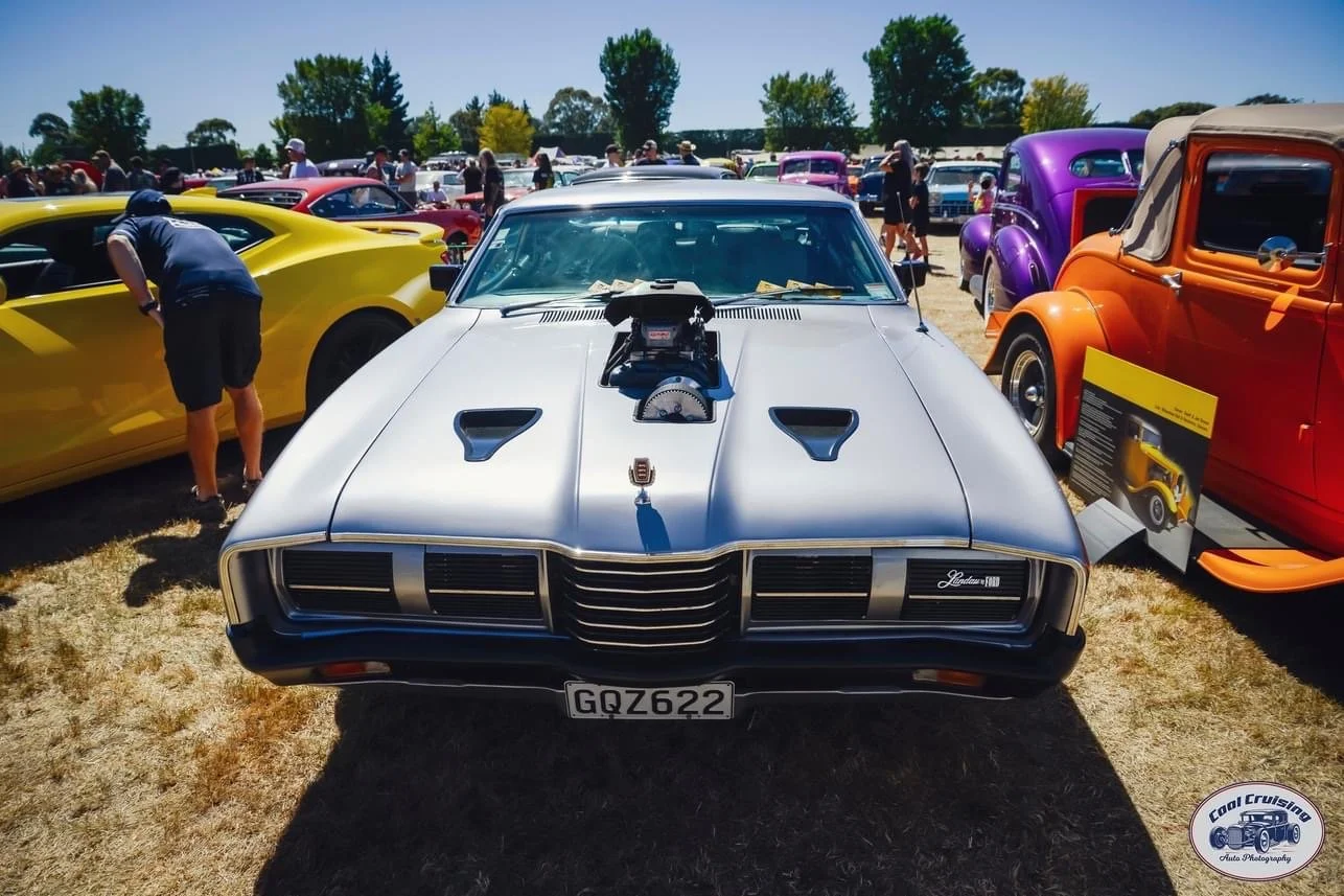 A silver classic Pontiac Firebird car with a supercharger on the hood at a car show, surrounded by various colorful vintage cars and spectators on a sunny day.