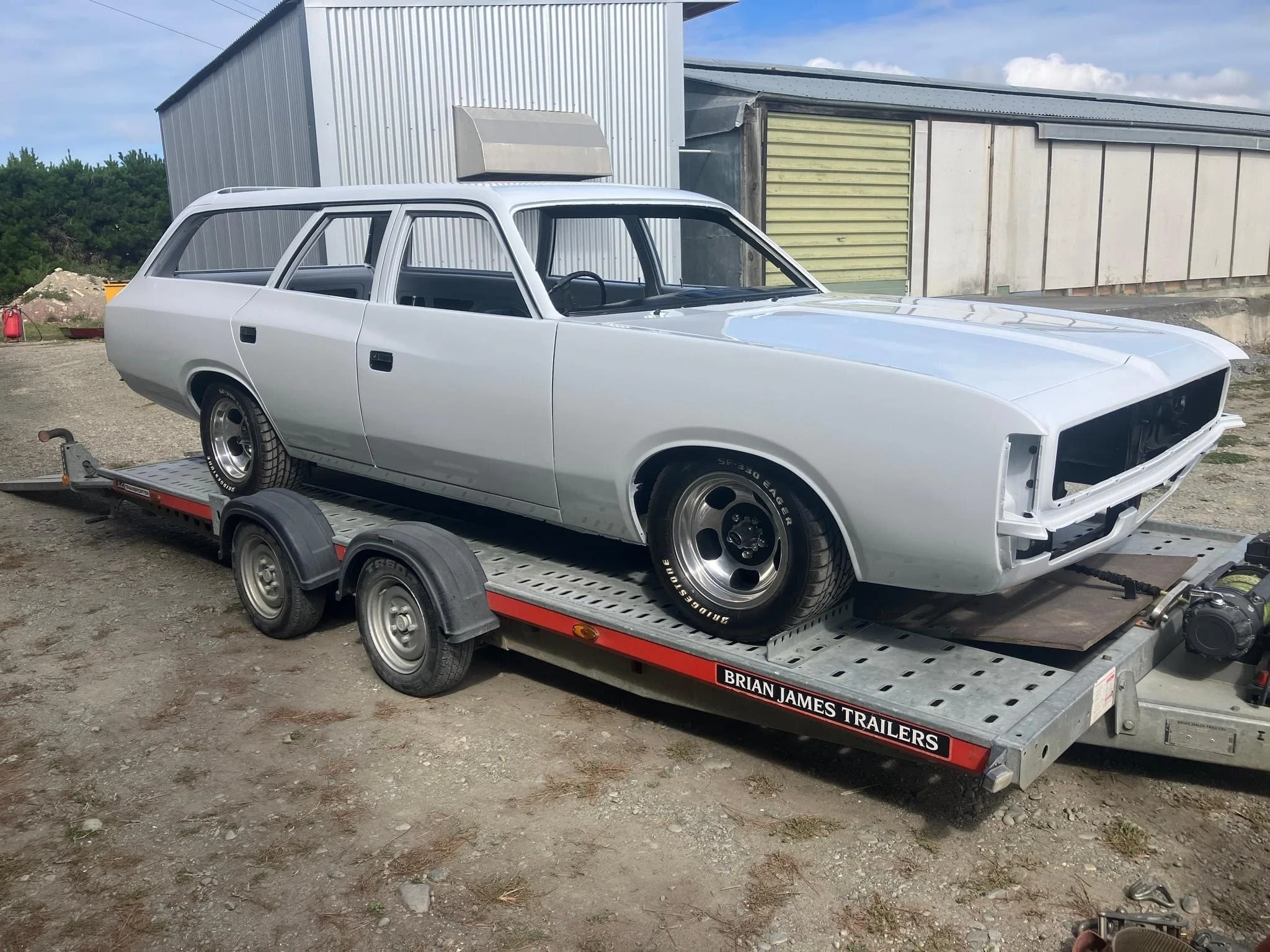 A white vintage car is on a trailer, appearing to be under restoration or repair, with no front grille or headlights installed. The trailer is parked outdoors on a gravel surface near a building with metal siding.
