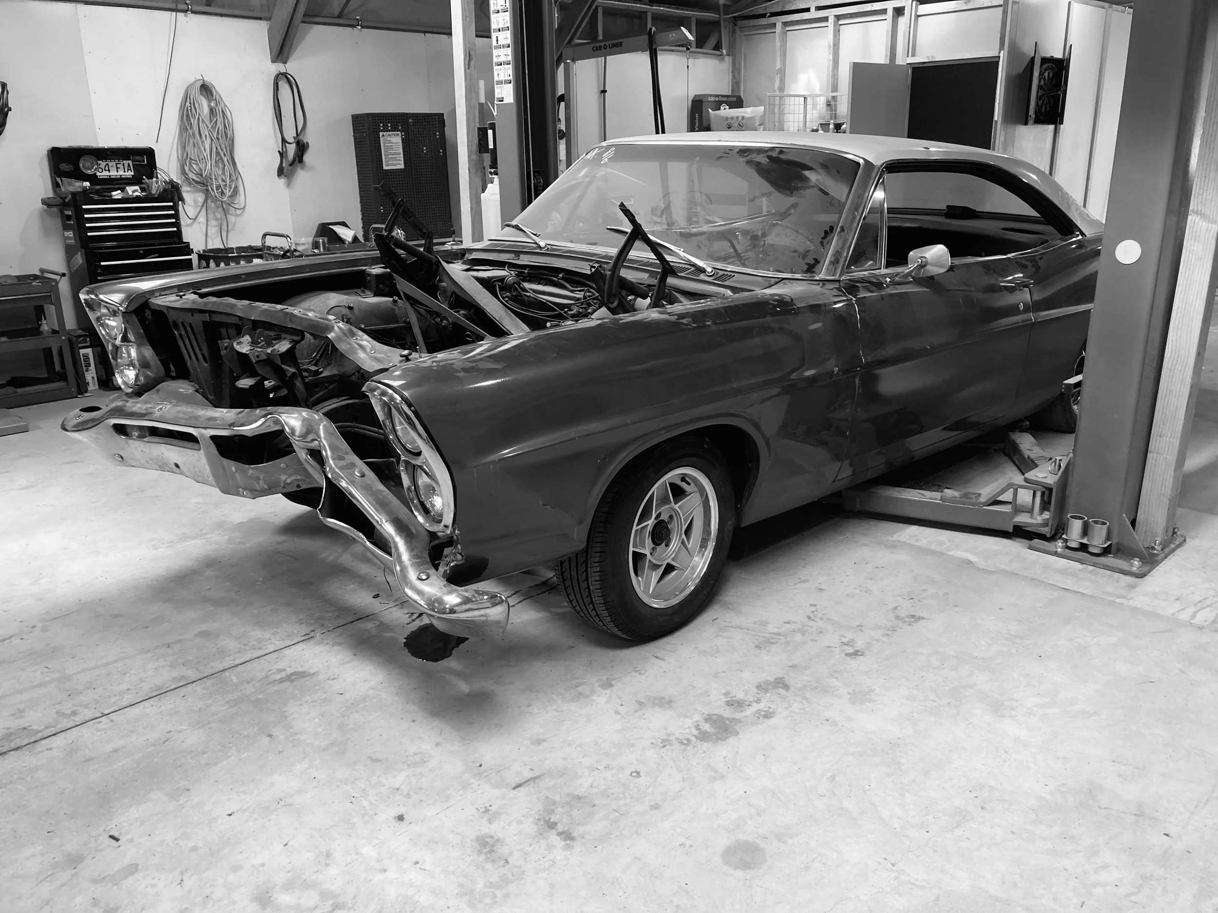 Black and white photo of a vintage car in a garage, with some parts removed, surrounded by tools and equipment.