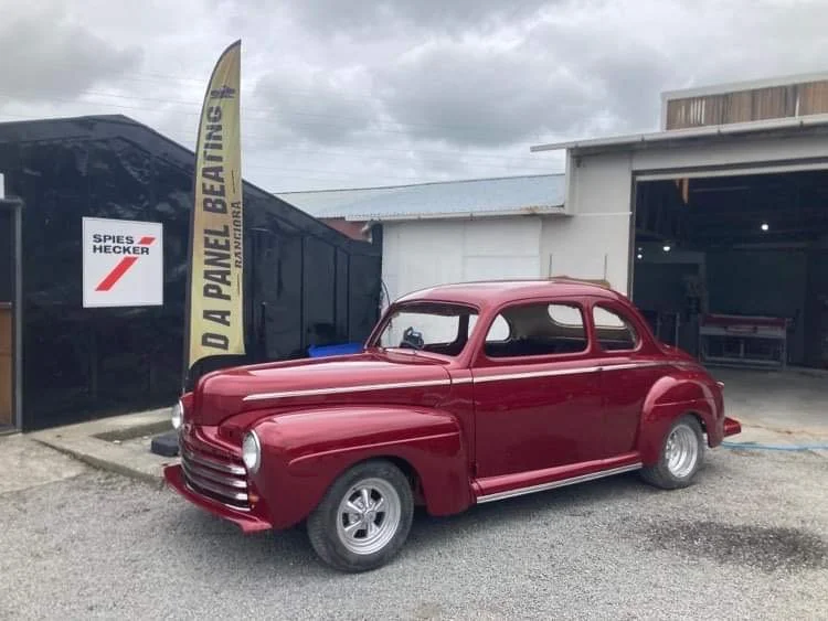 Red vintage car parked outside a building with a sign for 'Spies Hecker' and a yellow banner that says 'D A PANEL BEATING' on a cloudy day.
