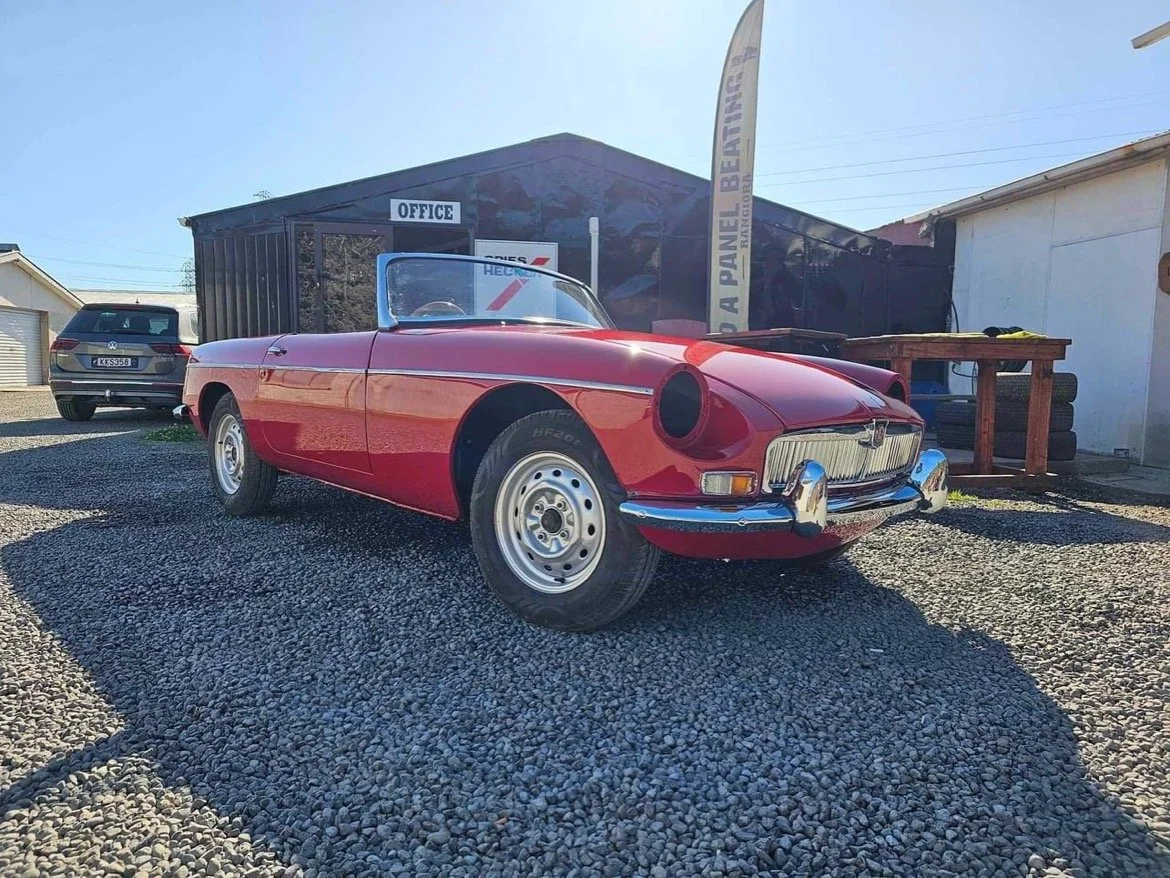 Red vintage convertible sports car parked on gravel outside a building with a sign for an office.