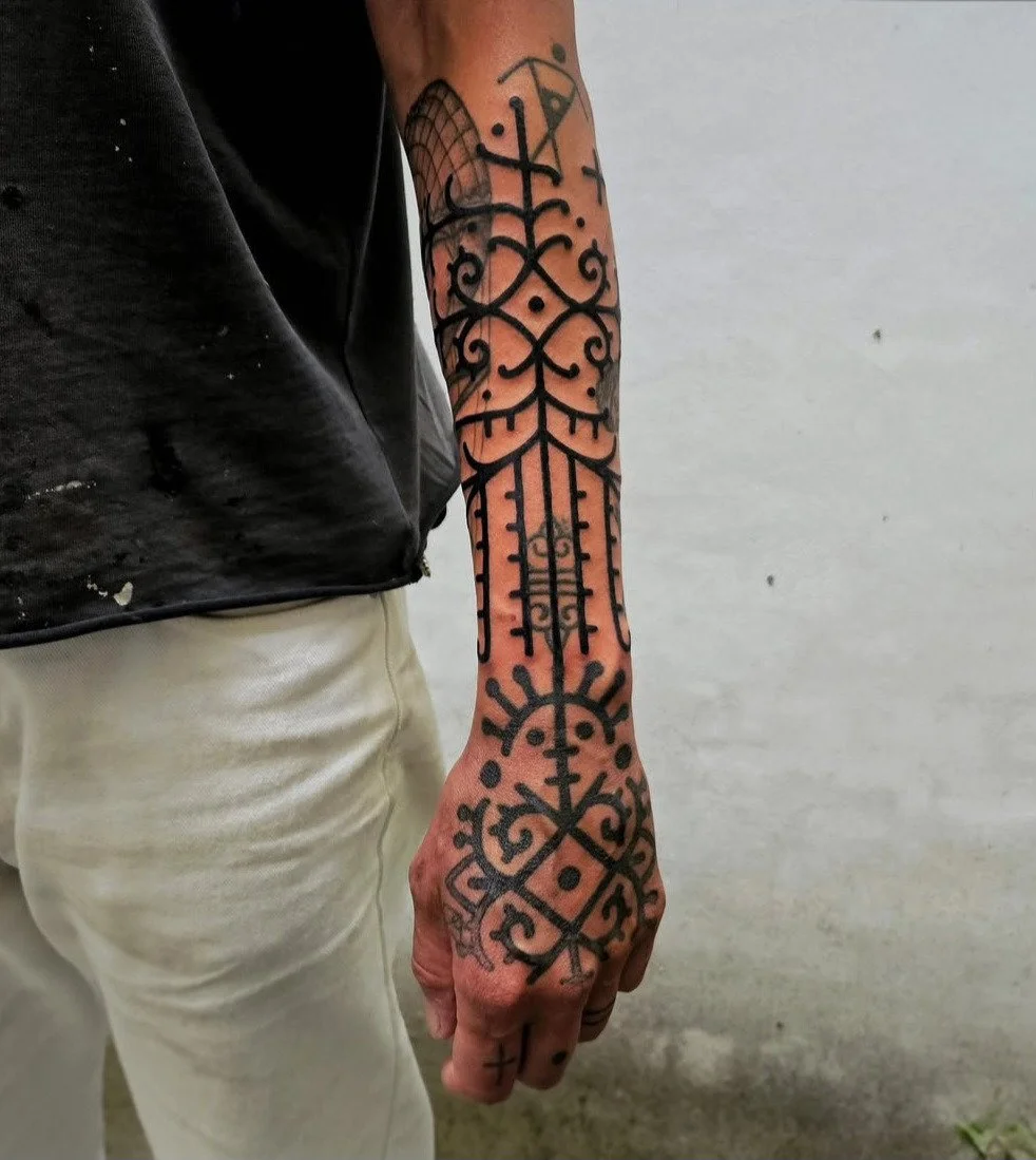 Een arm met een grote, zwarte, geometrische en figuurlijk getekende tattoo die zich uitstrekt van de pols tot ver op de bovenarm, met een grijze achtergrond en een persoon die een zwart shirt en lichte broek draagt.