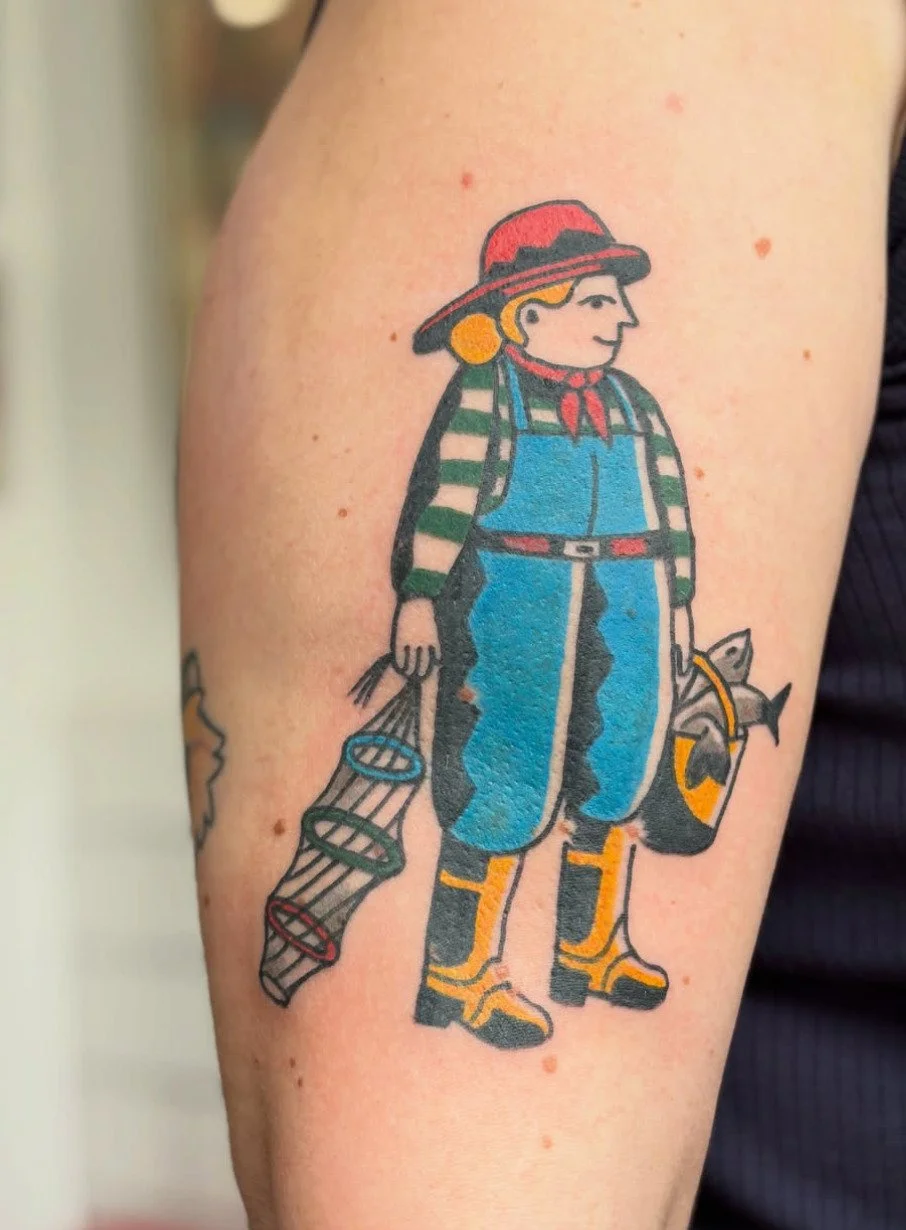 Tatoeage van een cartoonvissende jongen met een hoed, trui, blauwe spijkerbroek en rubberen laarzen, die een net en een vis vasthoudt.