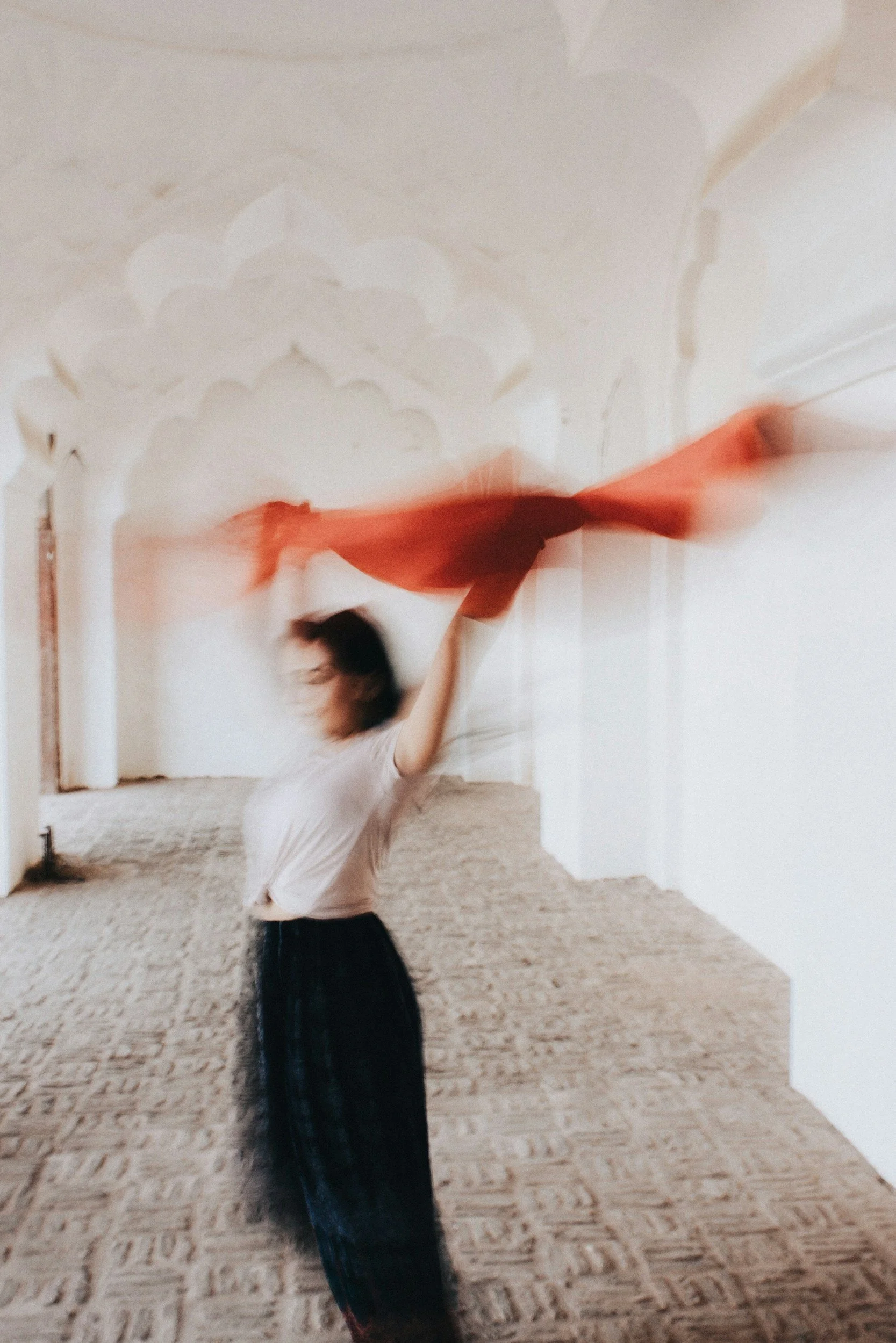 Blurred image of a woman with dark hair, wearing a white shirt and a black skirt, in a room with white walls and a stone floor, holding a red fabric or object that is moving.