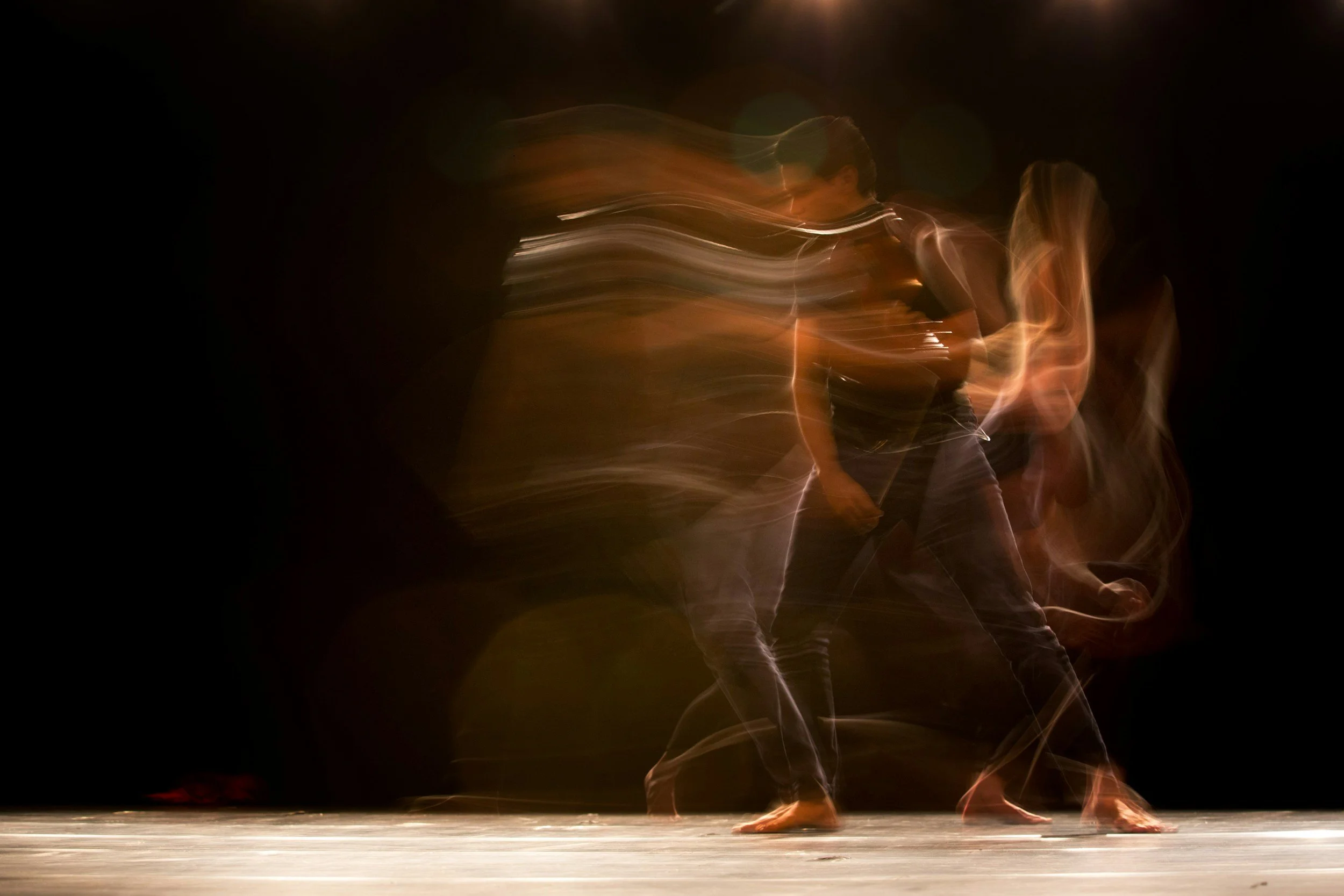 A dancer on stage with long hair and a black outfit, captured with motion blur against a dark background and a wooden floor.