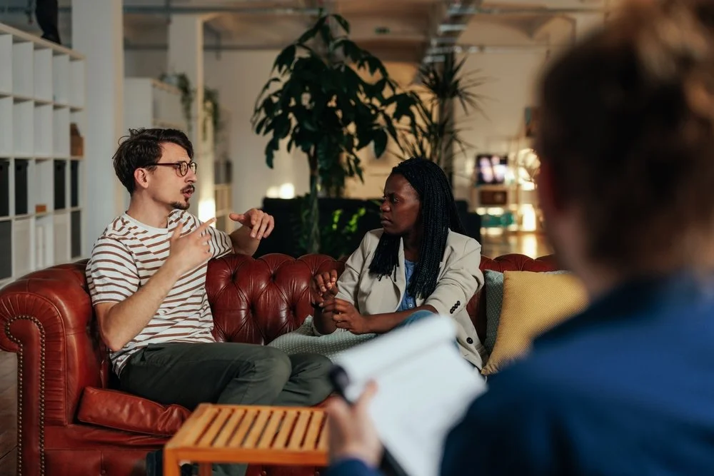 A man and woman sitting on a red leather couch engaged in a conversation, with another person in the foreground taking notes or asking questions, in a cozy, well-lit room with plants and bookshelves.