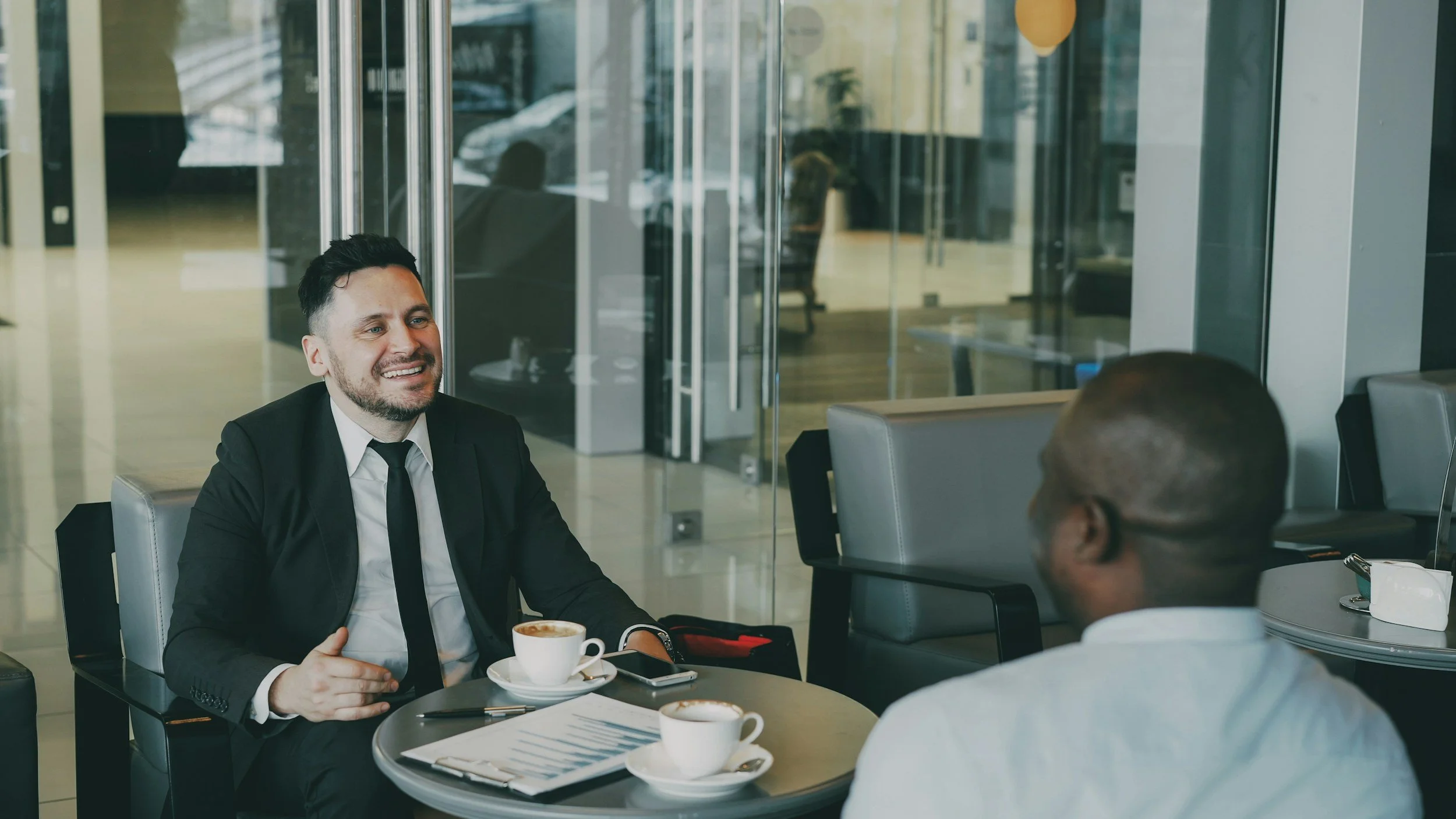 Two men having a business meeting in a café. One man is smiling, wearing a suit and tie, with a cup of coffee in front of him. The other man is partially visible. The café has glass walls and modern décor.