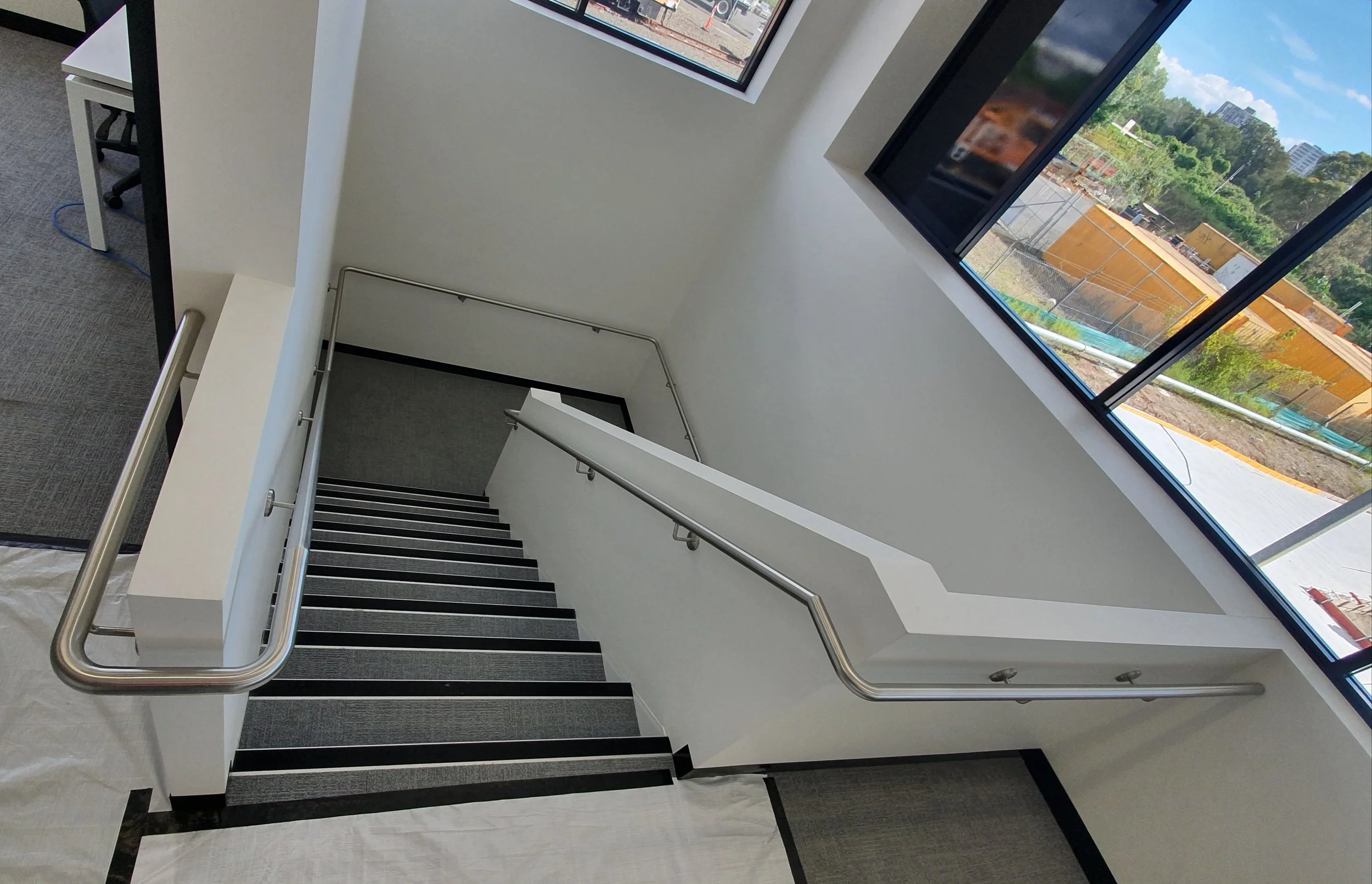 Interior view of a staircase with railings, lined with black and gray carpeting, adjacent to large windows that offer a view of an outdoor construction yard and trees.