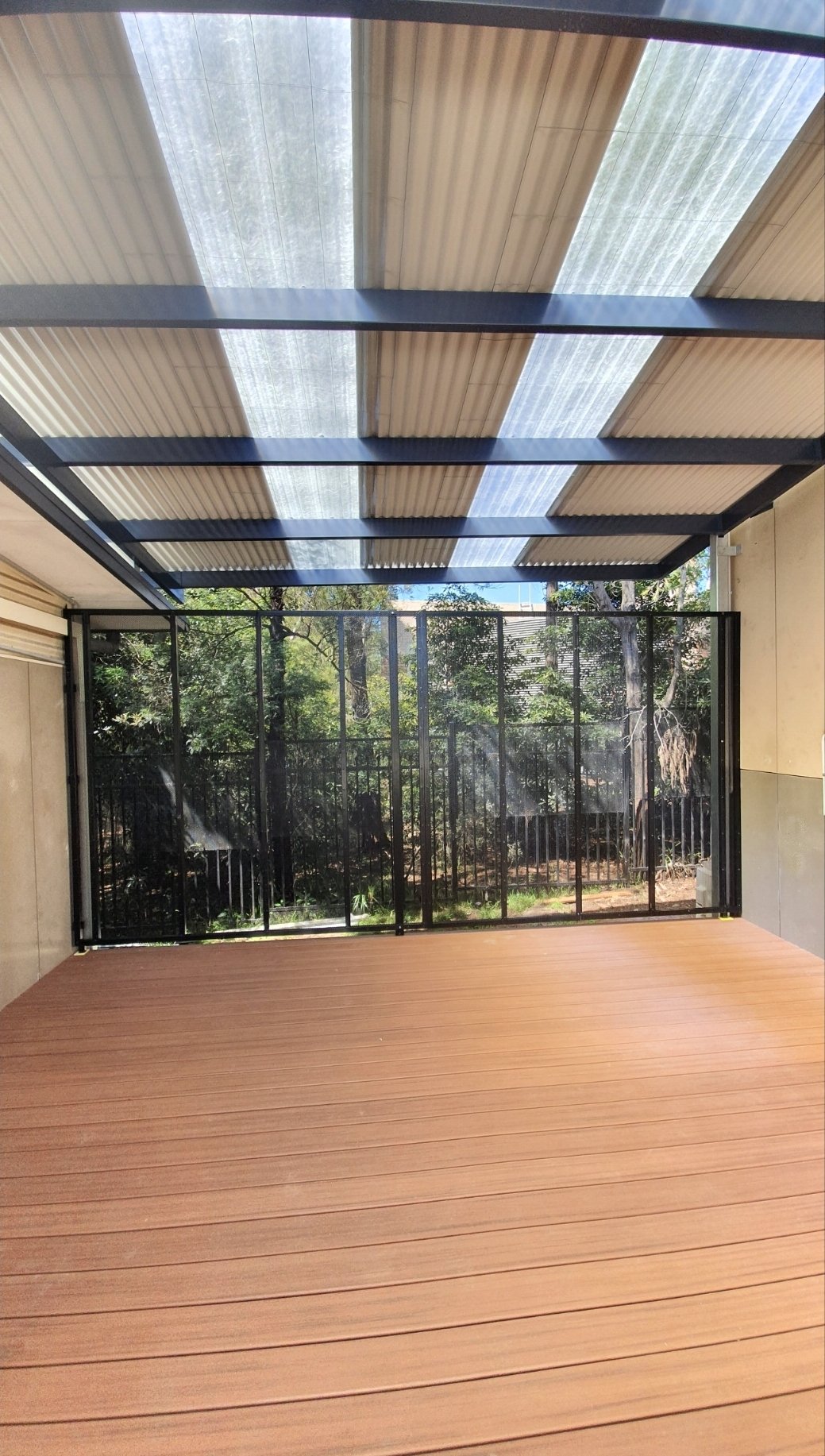 A balcony with a wooden floor, black metal railing, and a transparent roof with sections of frosted glass, overlooking a lush green outdoor area with trees.