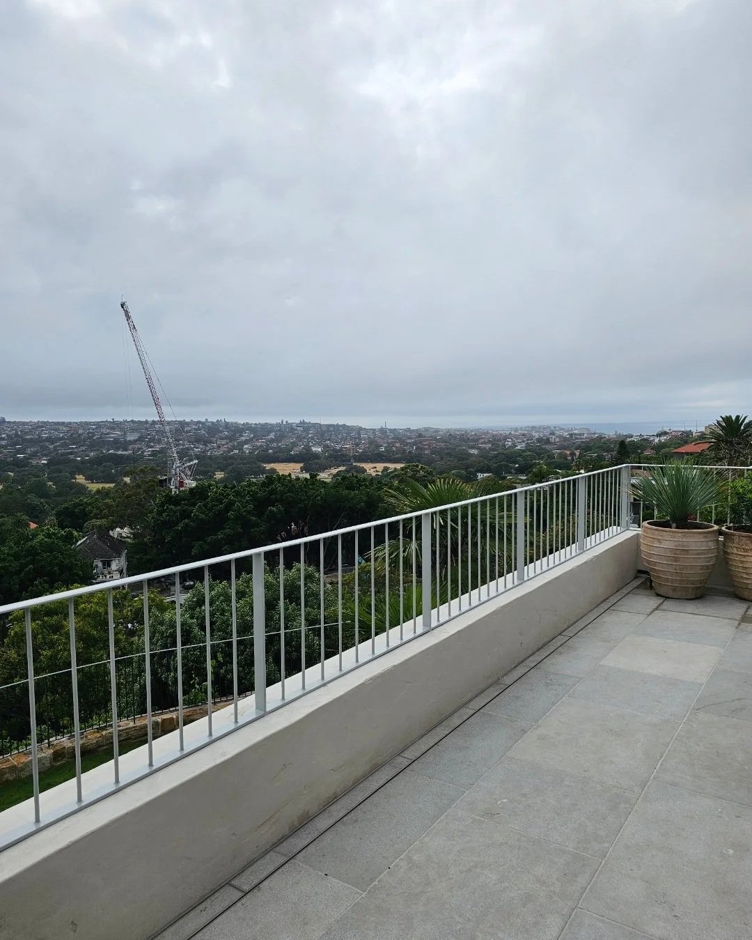 View from a balcony over a cityscape with trees, a tower crane, and a cloudy sky in the distance.