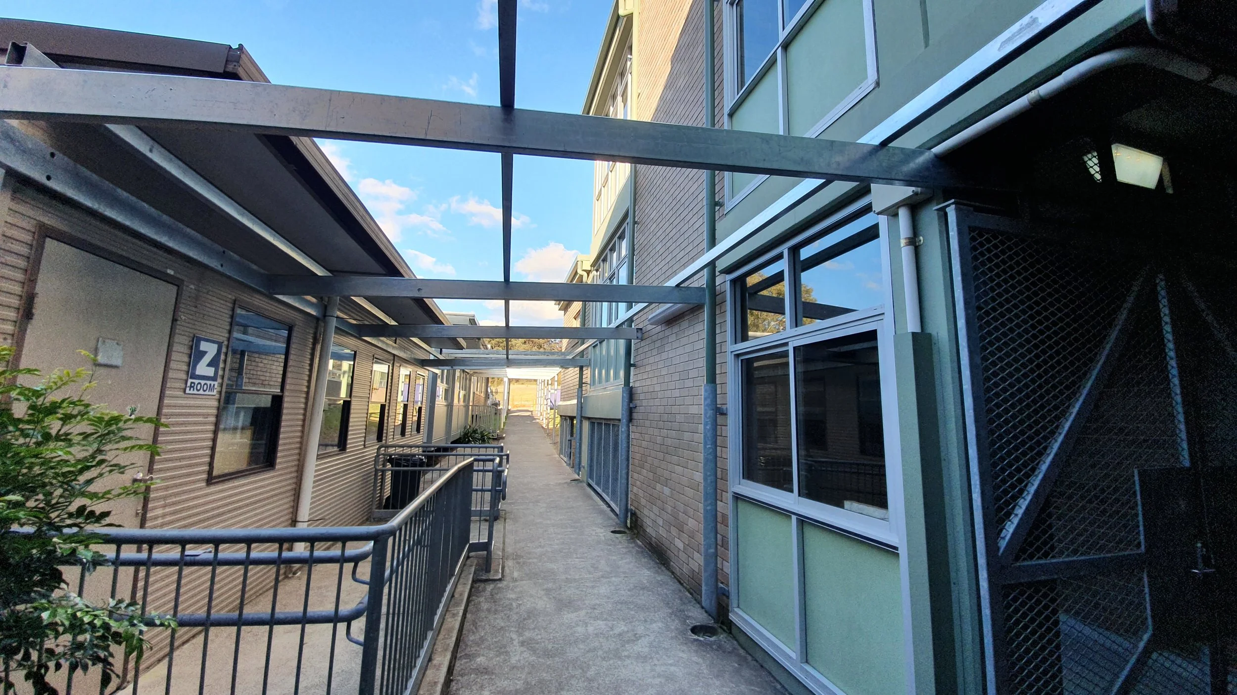 An outdoor walkway between two multi-story buildings with metal framework above, glass windows, and a railing along the pathway, under a partly cloudy blue sky.