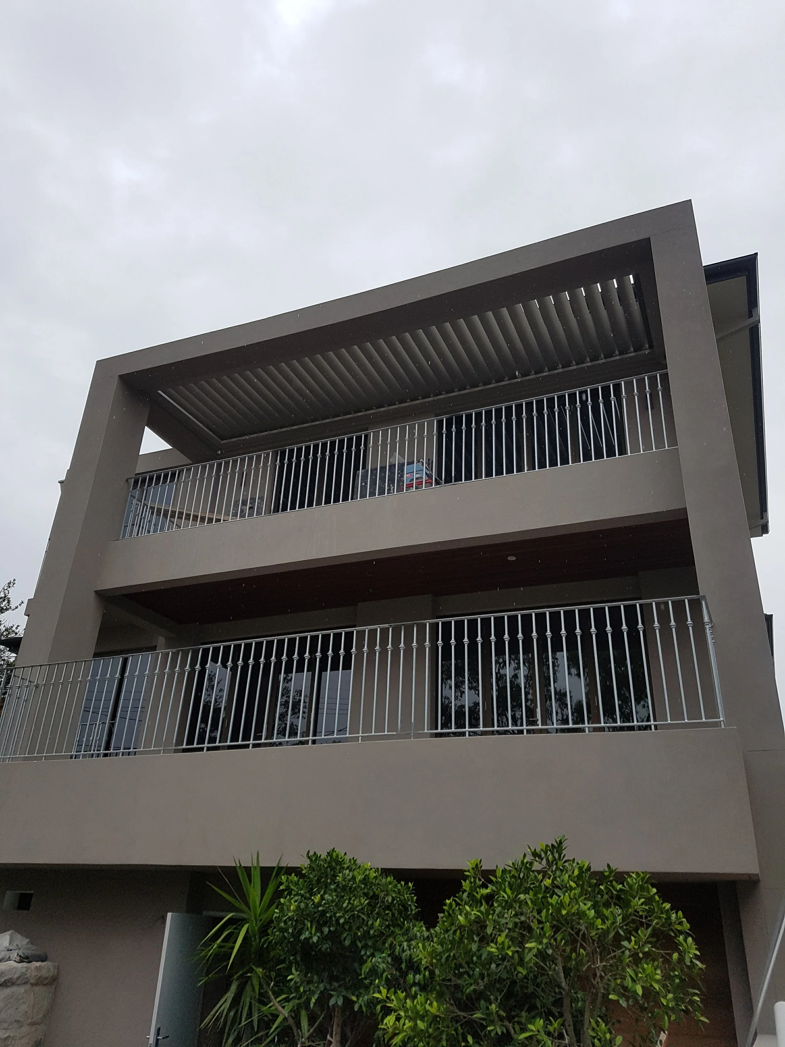 Modern multi-story building with balconies, metal railings, and a slatted roof over the upper balcony, with lush green bushes in the foreground and cloudy overcast sky.