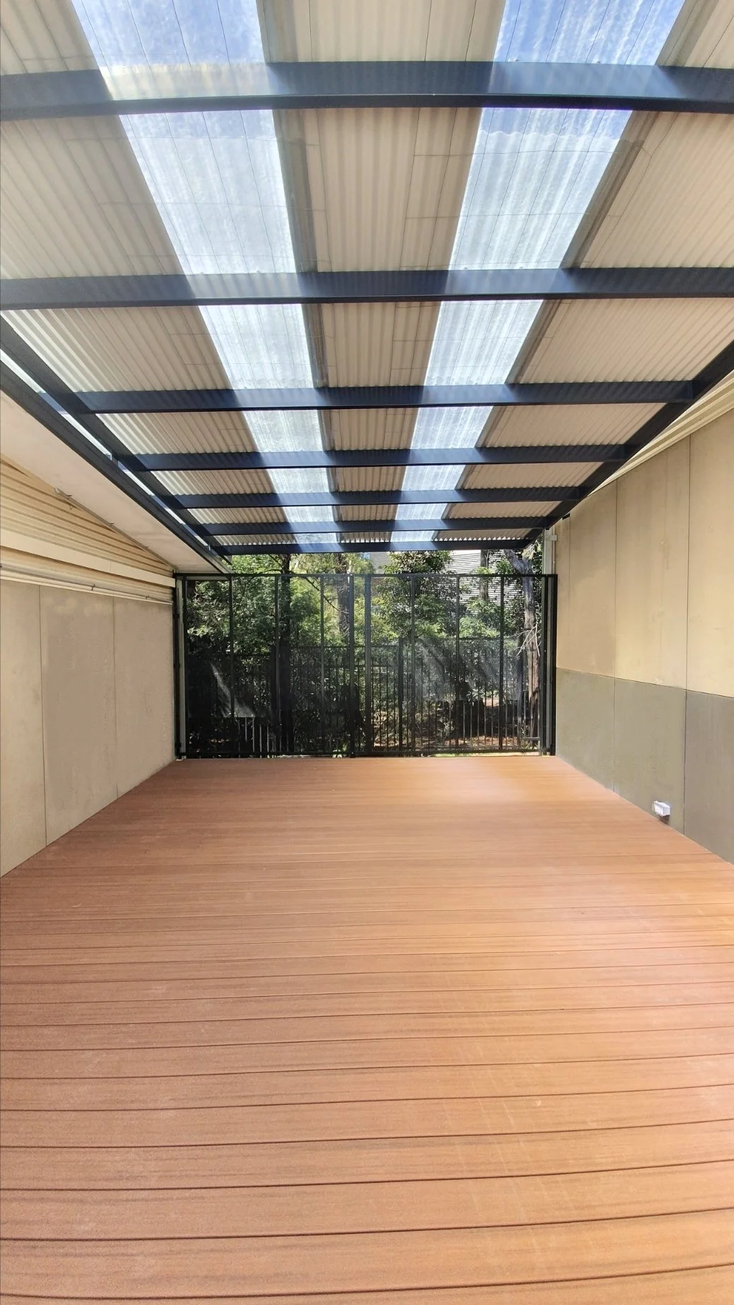Empty outdoor patio with wooden flooring, enclosed by metal and mesh fencing, with trees visible in the background and a partially transparent roof overhead.