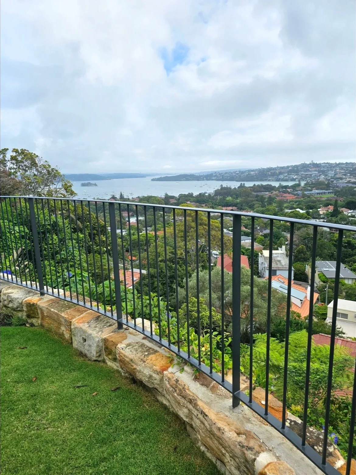 View of a city and waterbody from a balcony with black metal railing and green lawn in foreground.